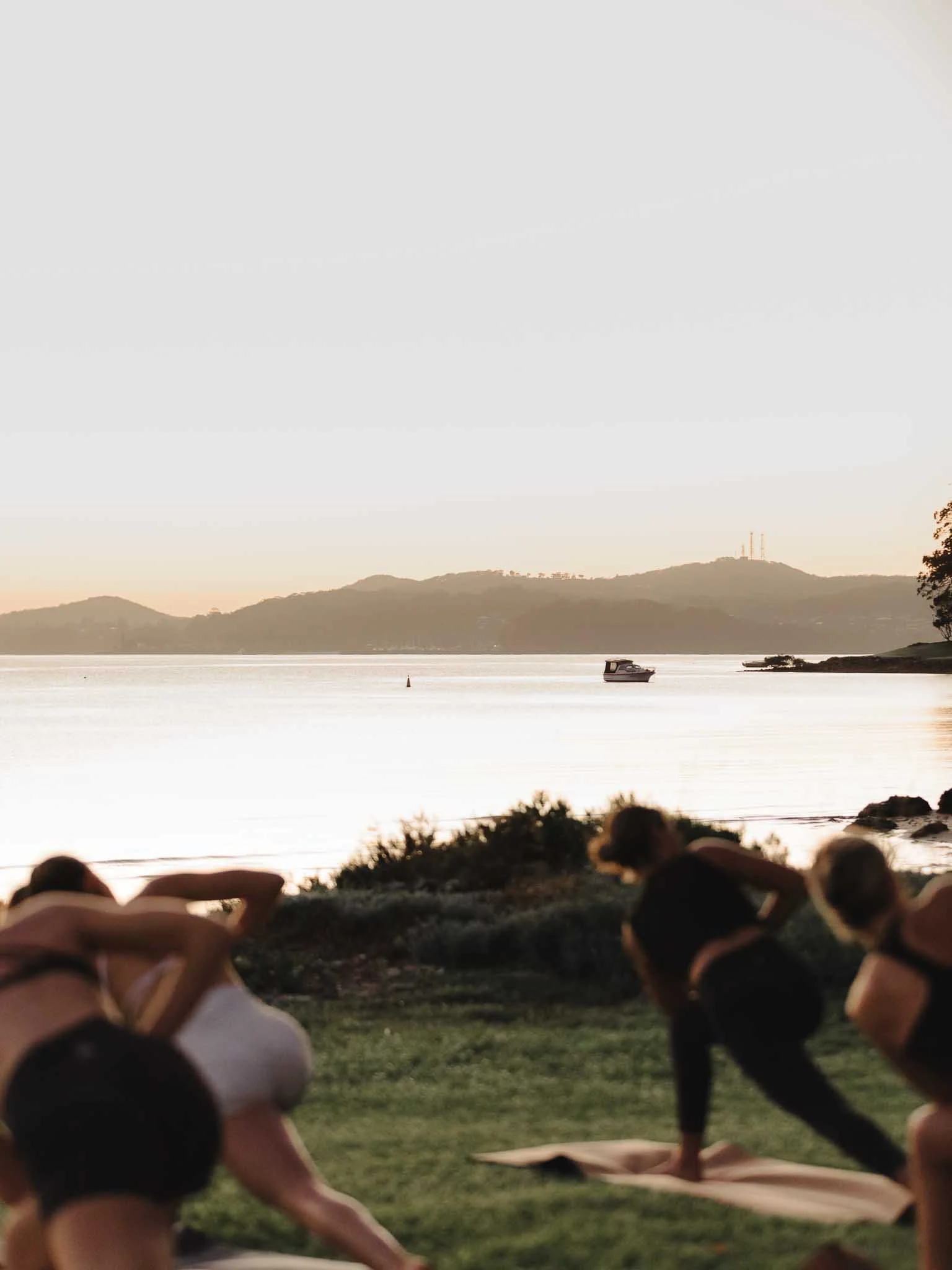 People doing yoga or stretching on mats outdoors near a body of water with hills in the background during sunset.