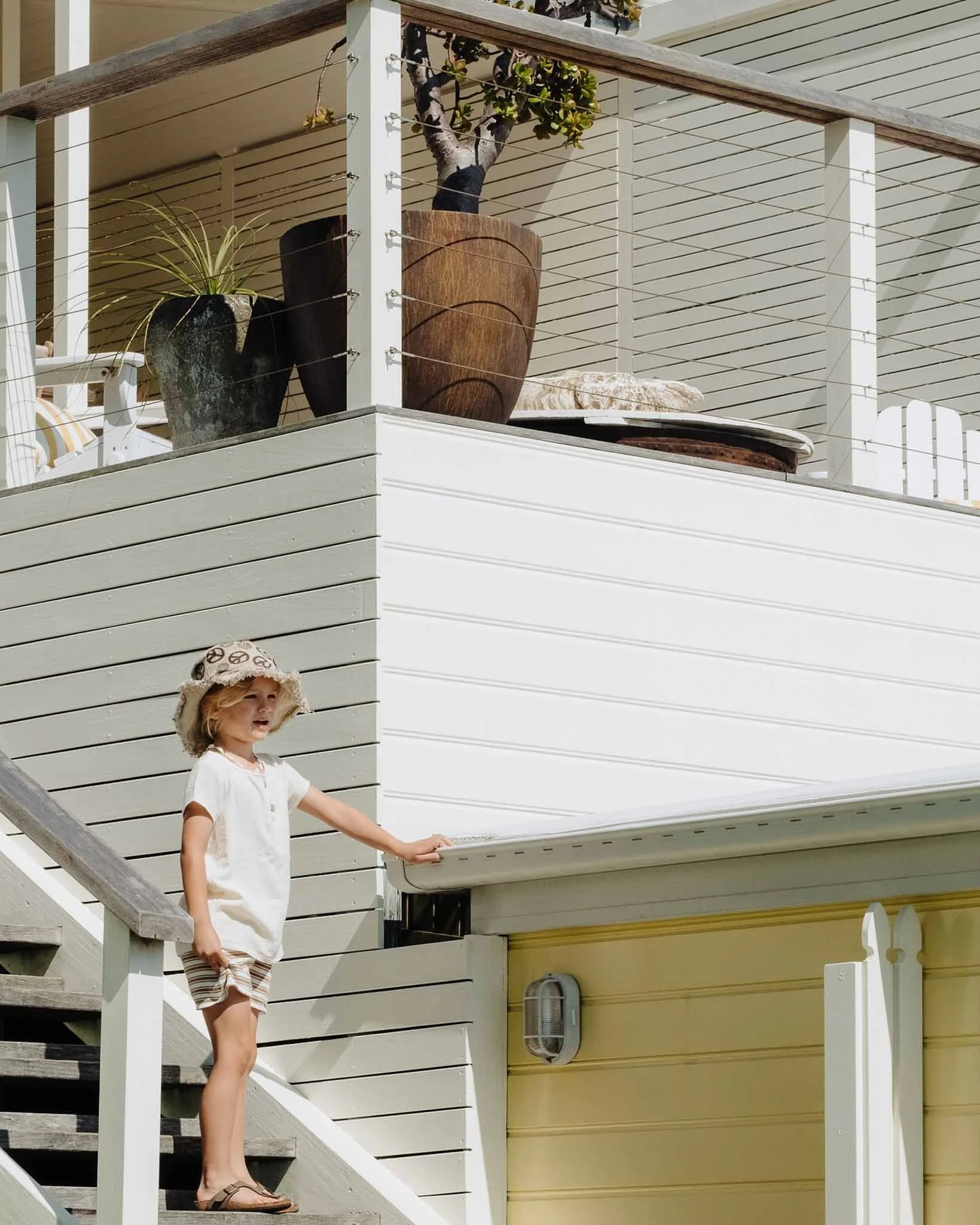 A young girl in striped shorts and a white shirt with a cream-colored hat stands on the stairs of a porch, holding a railing with her right hand, and looking off to the side. The porch has white siding with horizontal lines, a wooden railing, and potted plants including a small tree and a large decorative pot.