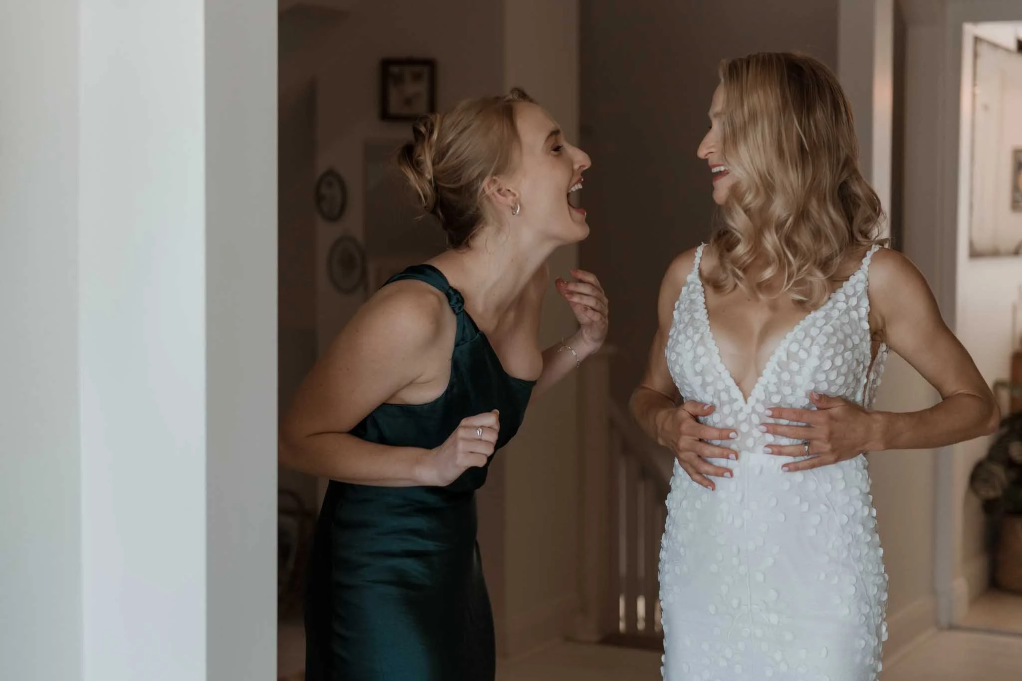 Two women, one in a wedding dress, laughing and talking indoors.
