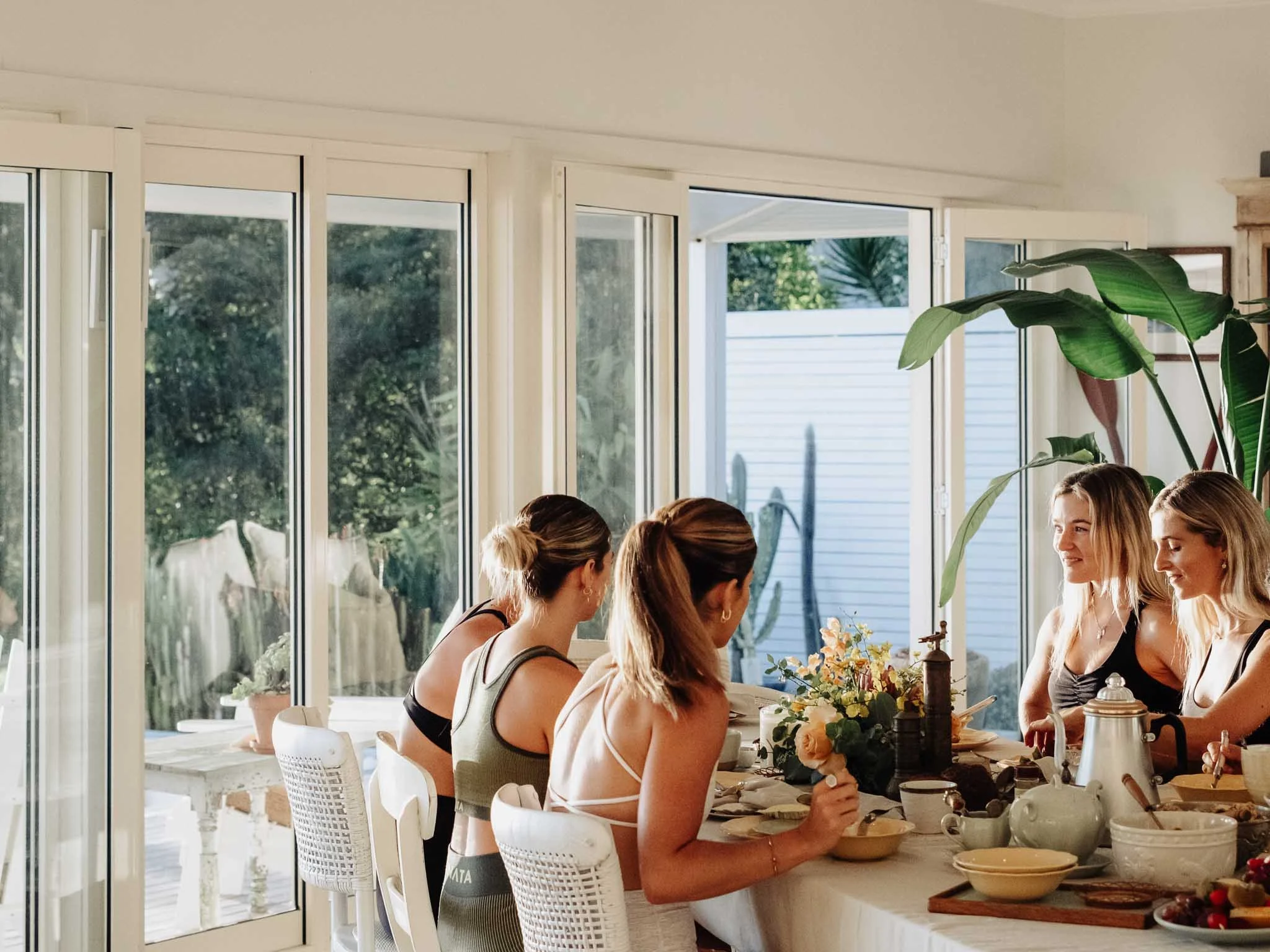 A group of women gathered around a kitchen counter, preparing food. Large windows with white frames behind them reveal a backyard with trees and a house. The kitchen has plants, flowers, and various kitchen utensils and dishes on the counter.