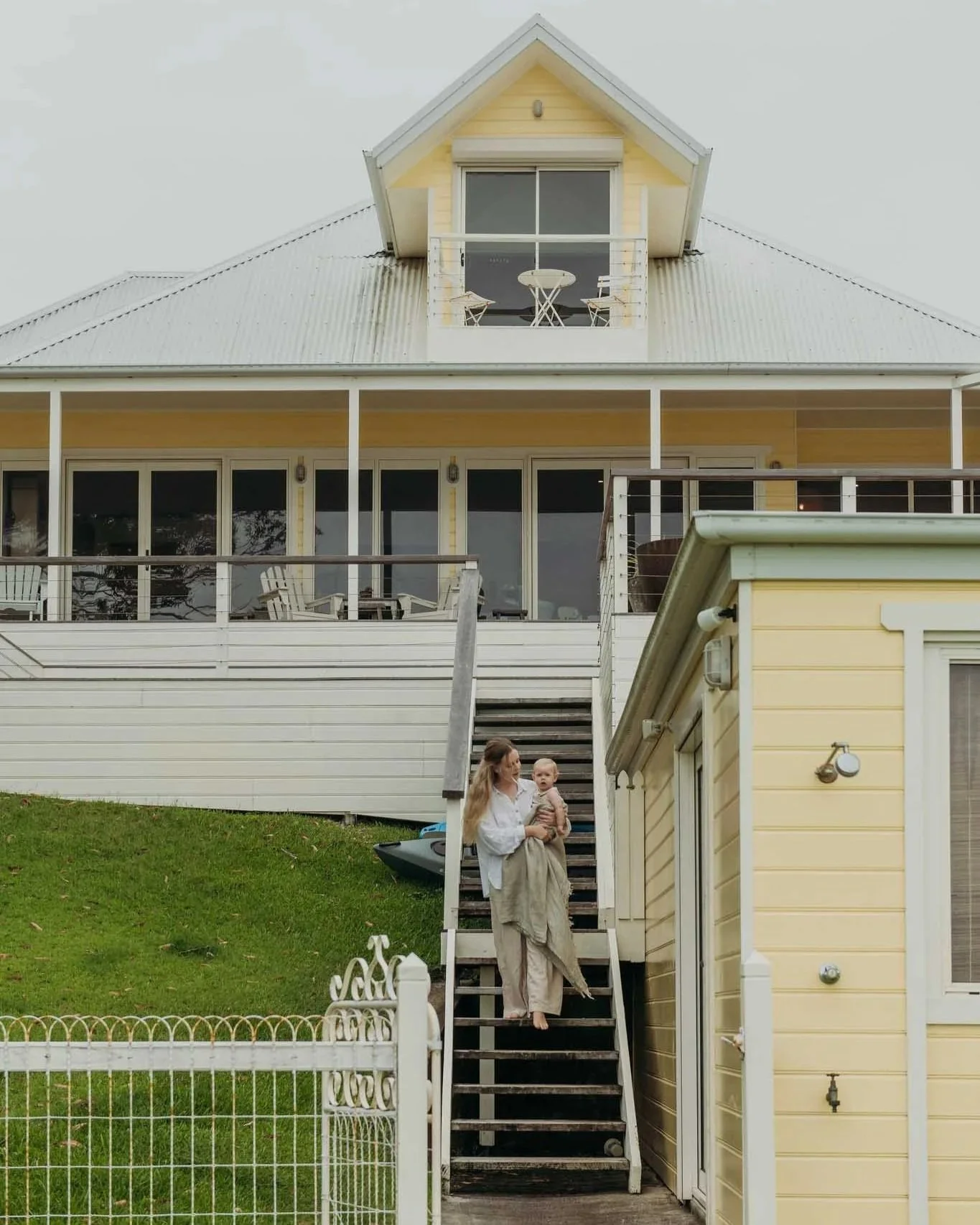 A woman holding a baby standing on an outdoor staircase in front of a yellow house with large glass doors and a balcony.