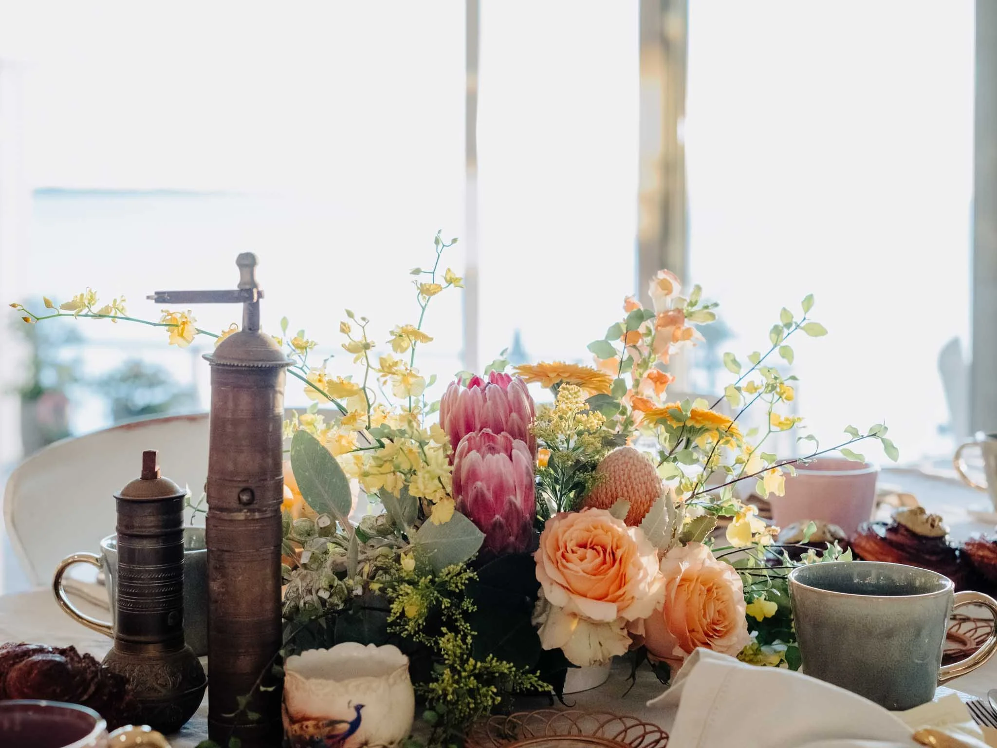 A table with a floral centerpiece featuring various colorful flowers, a small rustic coffee grinder, and teacups. The background shows bright natural light coming from a window.