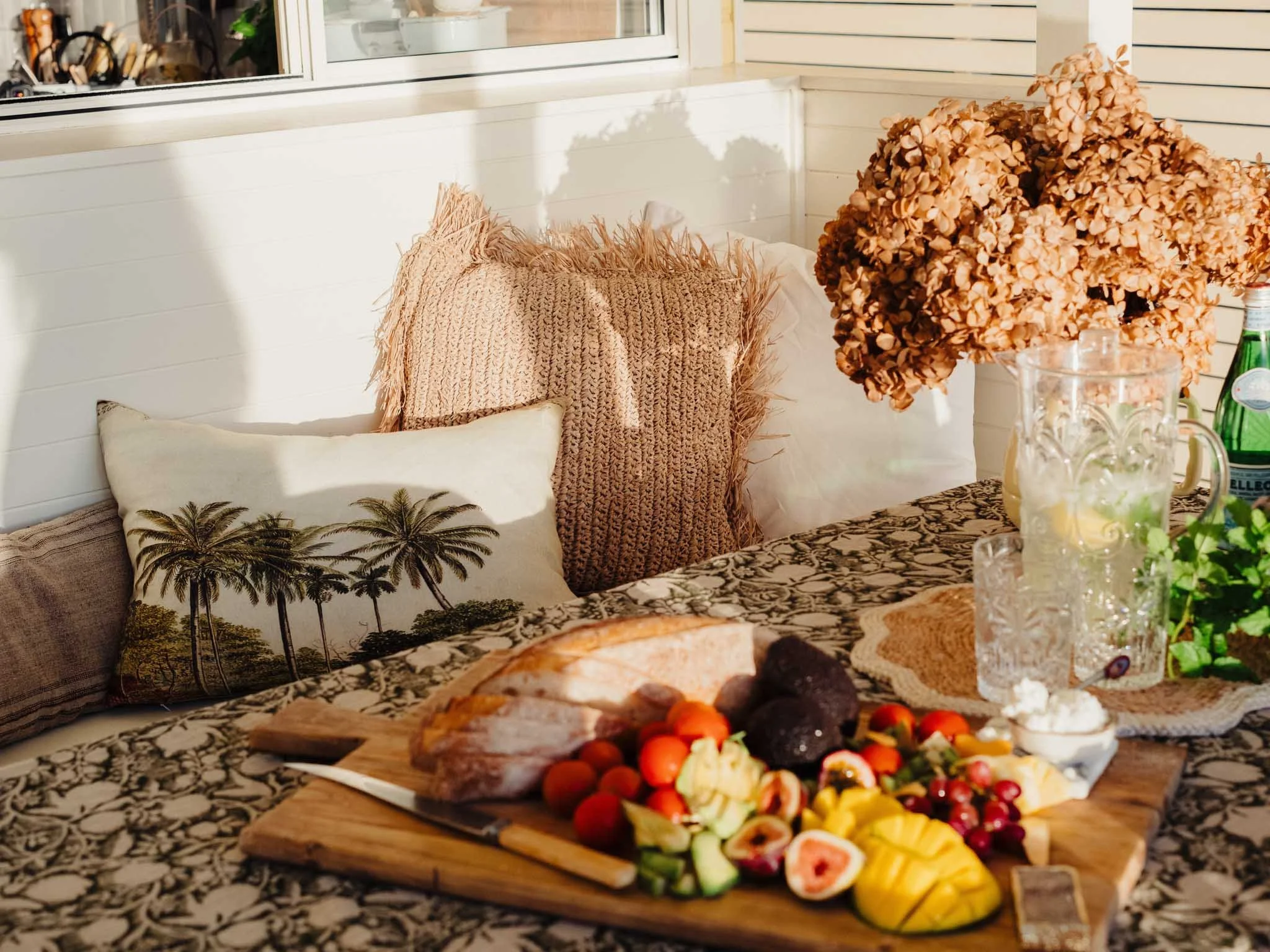 A wooden table with a tray of sliced fruits, including strawberries, grapes, pineapple, and kiwi, on a patterned tablecloth. Behind the tray are glass pitchers with drinks and a large bouquet of dried hydrangeas.