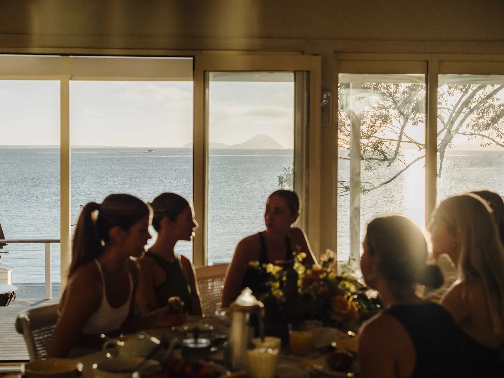 People dining indoors near large windows with a view of the ocean, trees, and mountains in the background, during sunset.