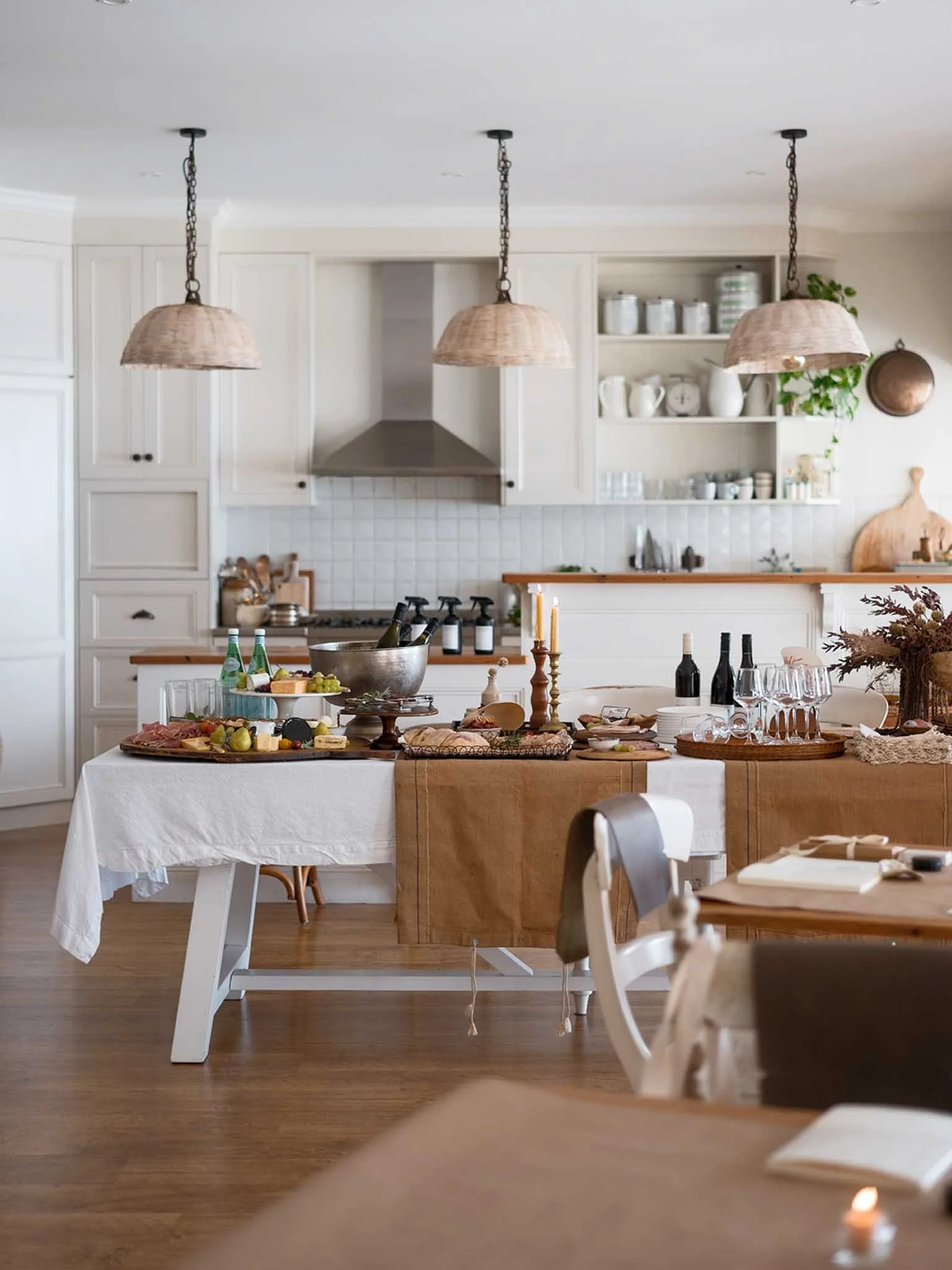 A kitchen decorated for a meal with a table set with foods, drinks, and candles, white cabinets, open shelves with dishes and jars, wooden accents, and hanging pendant lights.