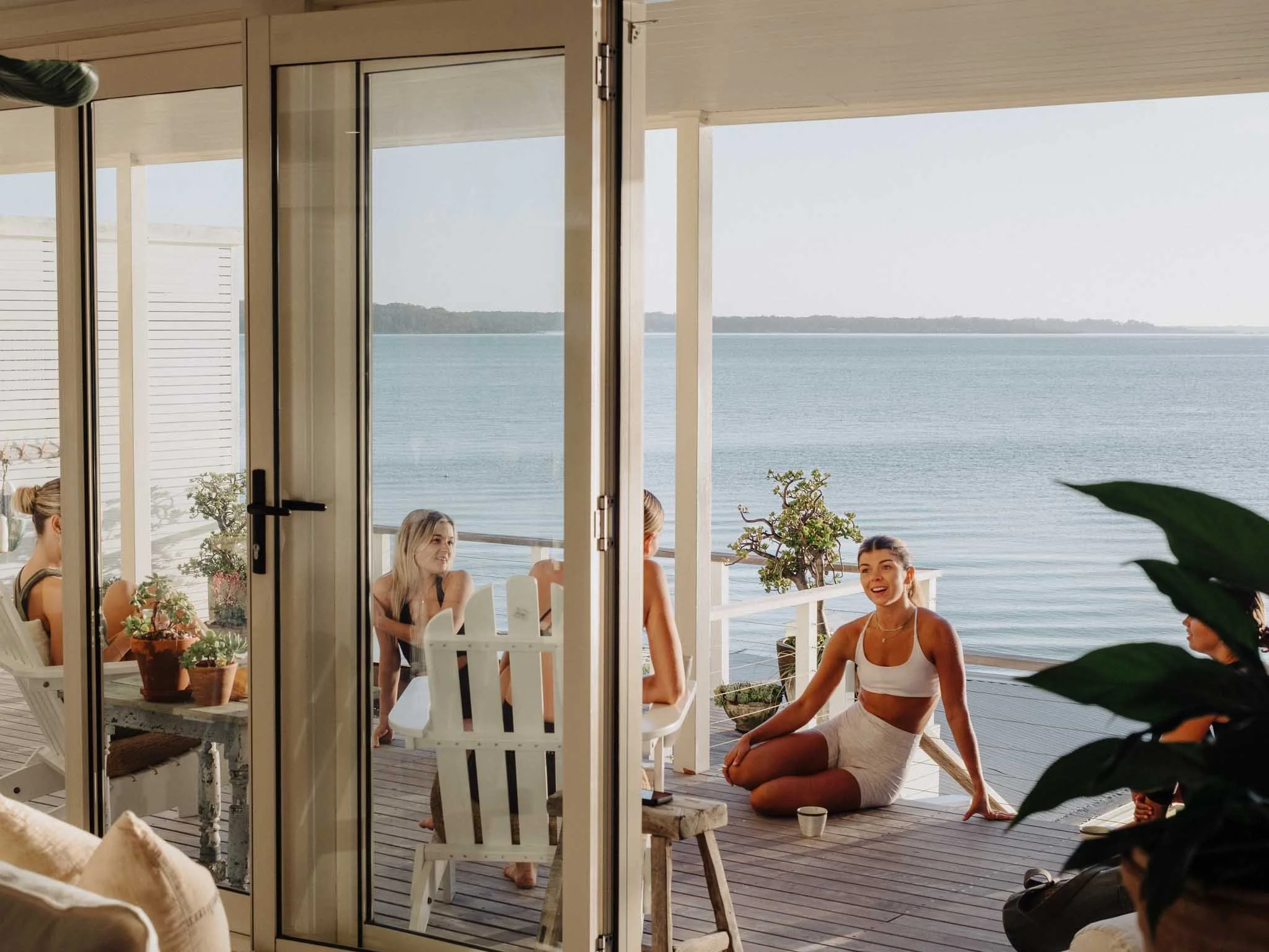 People sitting and kneeling on a wooden deck outside by the water, seen through a framed glass door, with some potted plants on a table inside.
