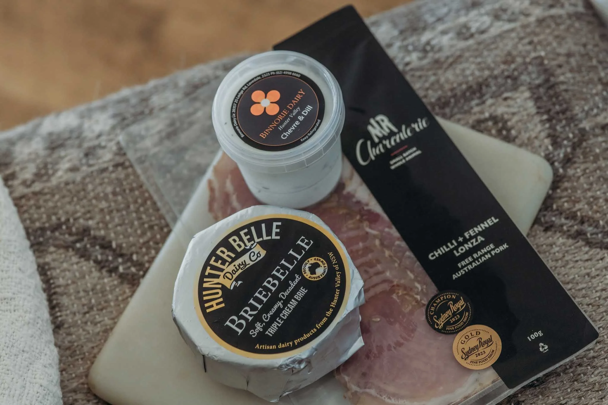 A tub of Hunter Valley triple cream Brie cheese, a package of Hunter Valley chicken and fennel loin, and a small jar of Binorrie Dairy butter on a beige surface.