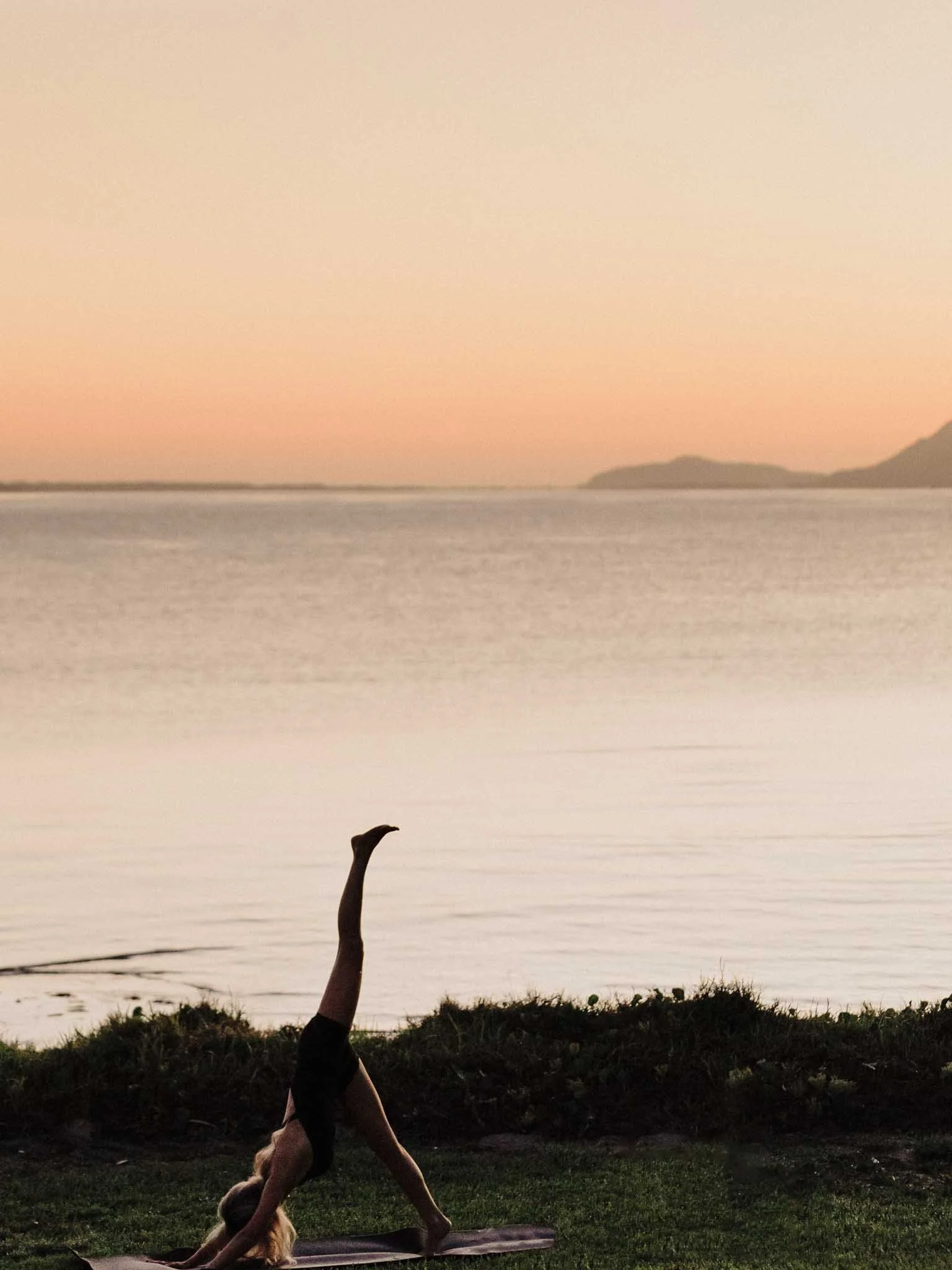 A dog practicing yoga outdoors at sunset, doing a handstand on a yoga mat with a body of water and hills in the background.