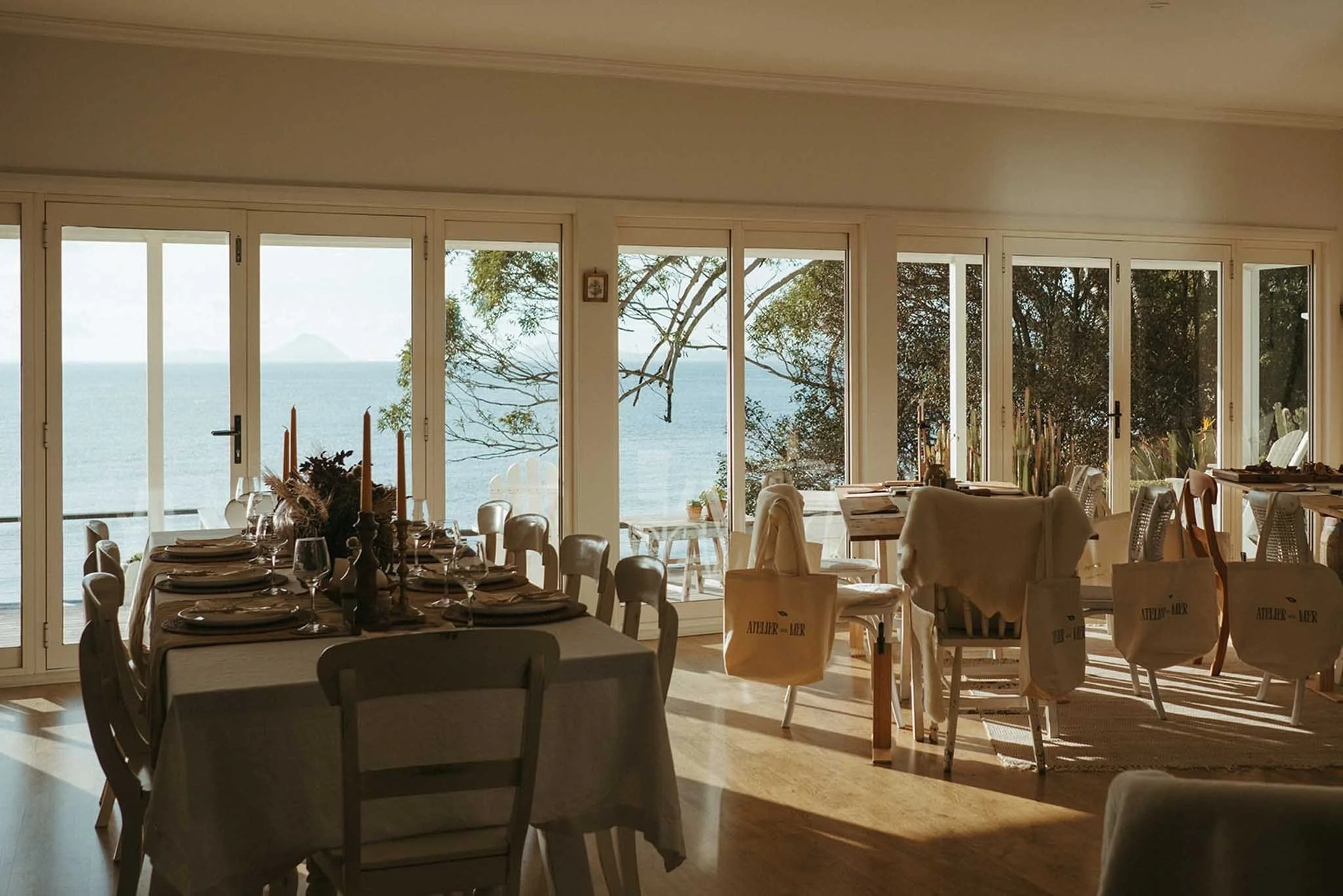 Dining area with tables set for a meal, large glass windows overlooking the ocean with trees outside, and sunlight casting shadows inside.