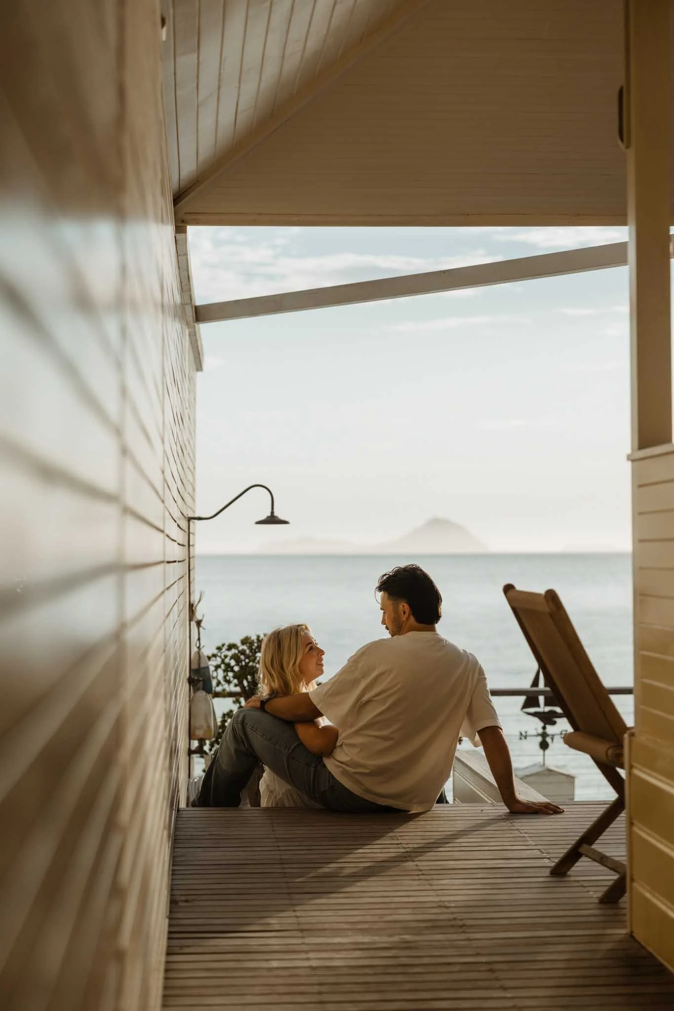 A couple sitting closely on a balcony or deck, facing each other with ocean and distant mountain view in the background, sharing an intimate moment during sunset.