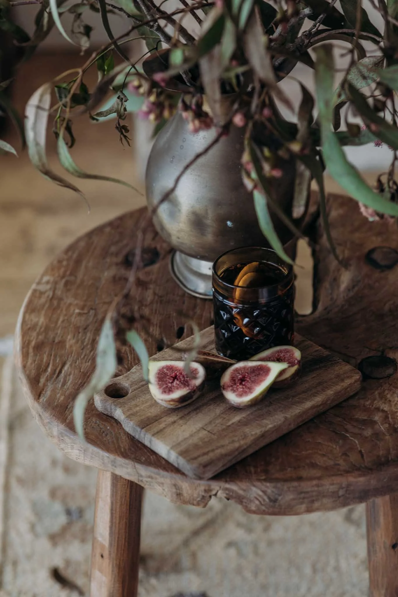 Close-up of a rustic wooden table with a wooden cutting board on top. Sliced figs with pink insides are arranged on the cutting board. A dark-colored glass with a drink and a silver vase with dried, twisted, and green leaves and flowers are also on the table. The setting has a cozy, natural aesthetic.