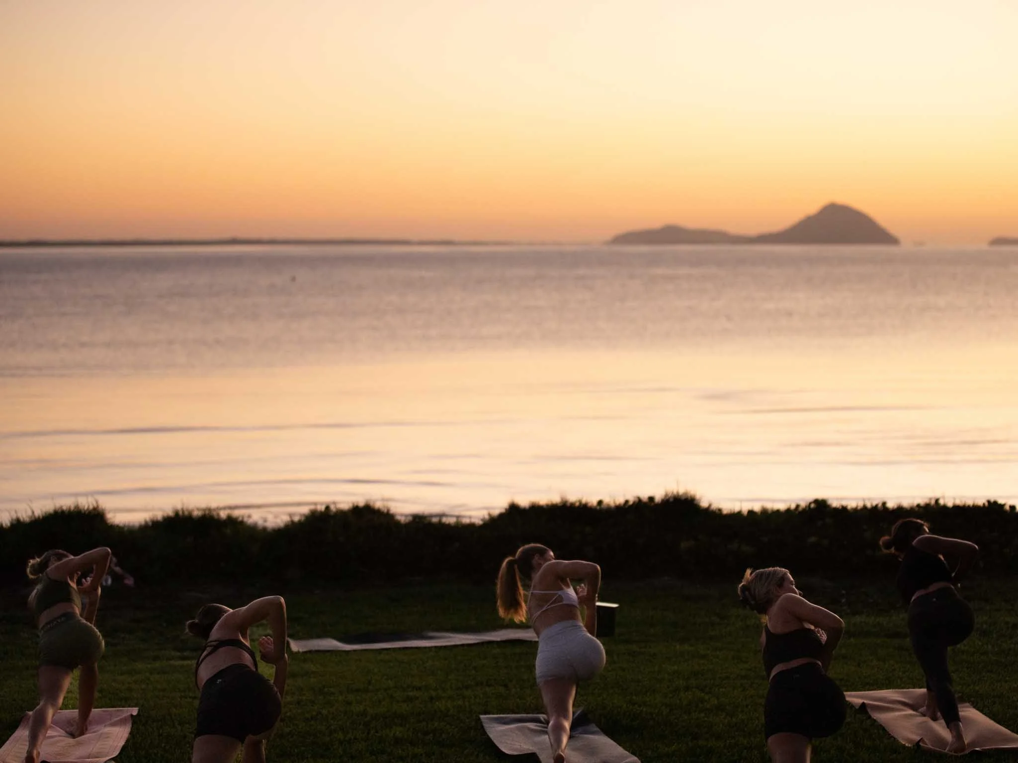 Group of people practicing yoga outdoors at sunset, near a body of water with distant mountains.