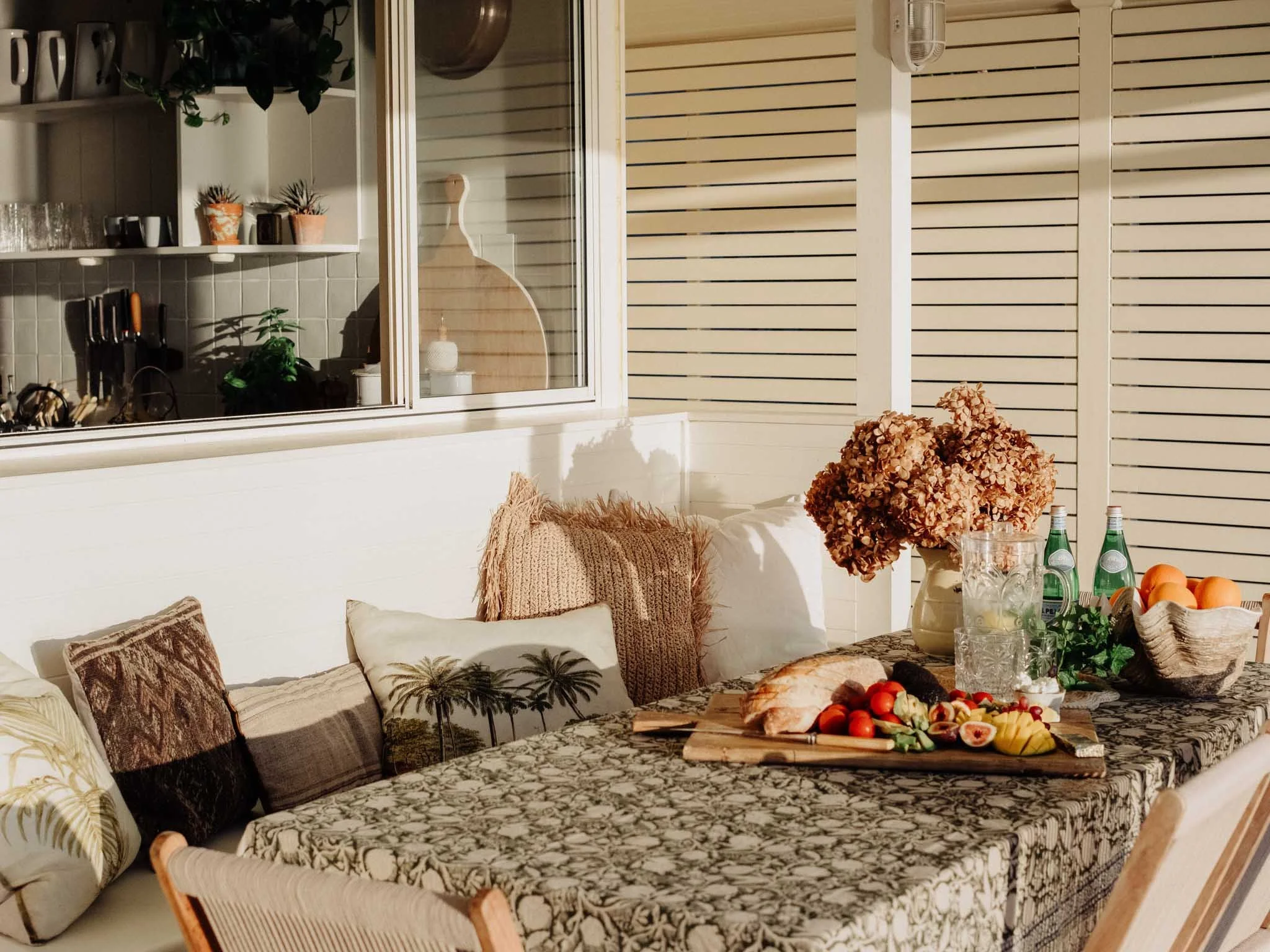 A cozy indoor dining area featuring a marble patterned tablecloth, assorted pillows on a white cushioned bench, a bouquet of dried flowers, and a spread of fresh food including tomatoes, cheese, and bread. The background shows a white paneled wall, a