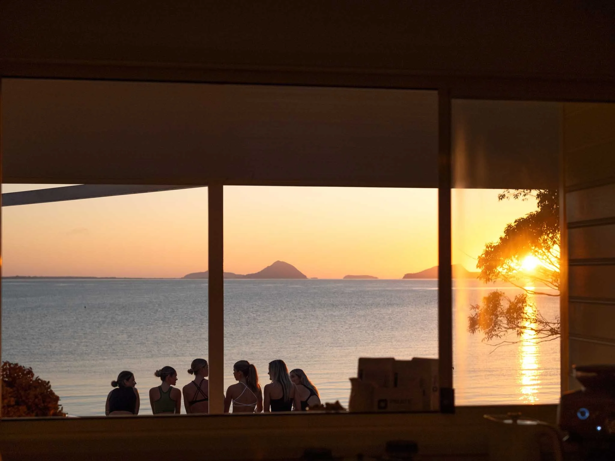 View of the ocean at sunset with distant islands, seen through a window with reflections and silhouettes of five women standing and talking.