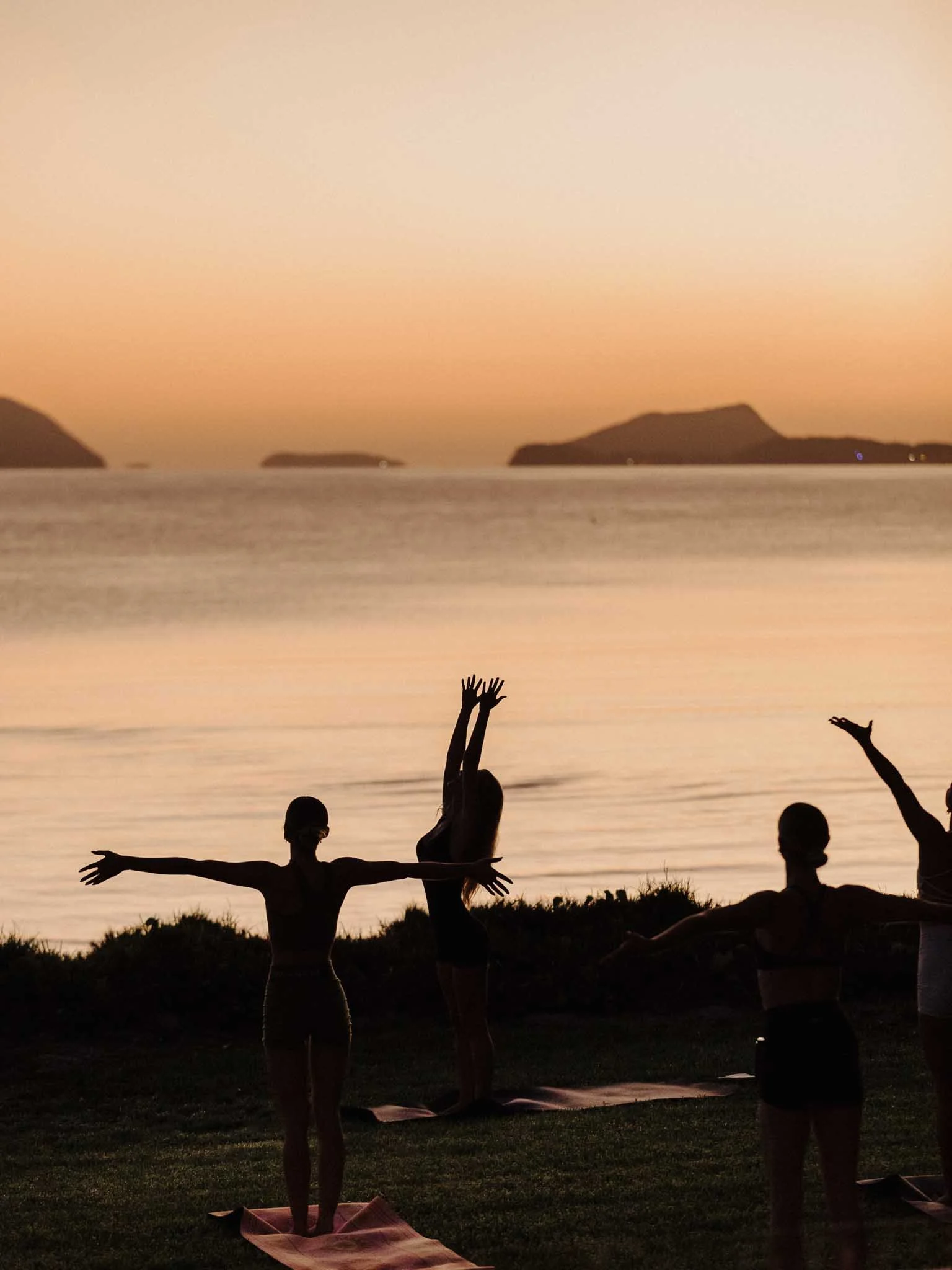Group of people doing yoga on mats by the water during sunset with islands in the background.