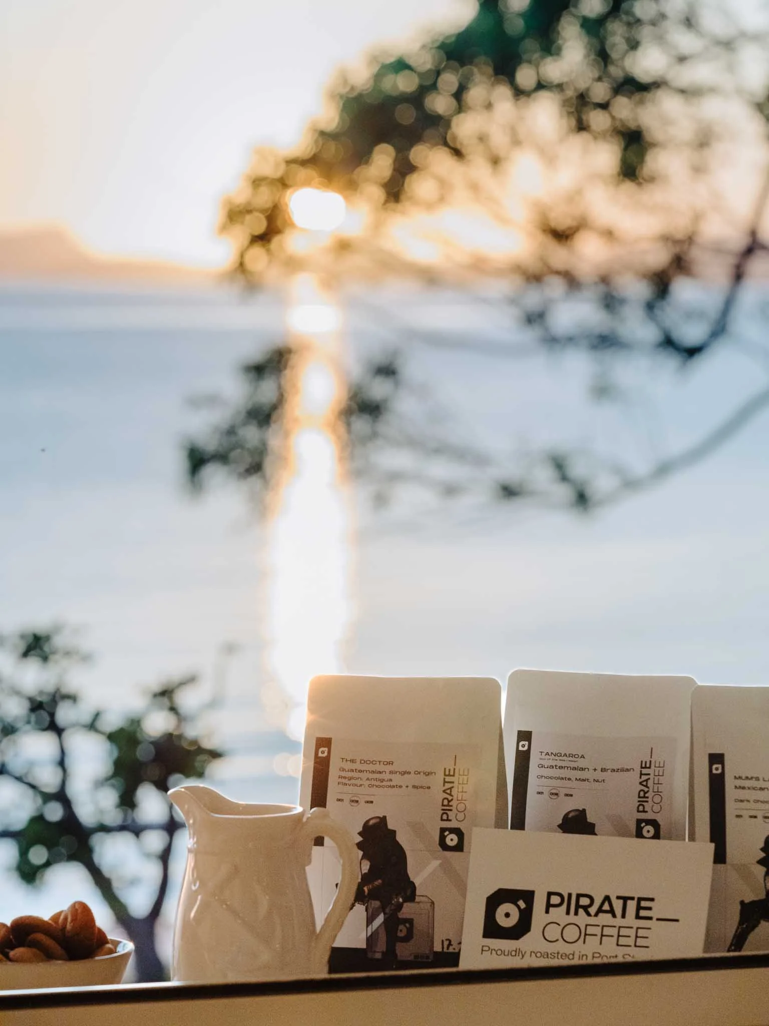 Several packets of Pirate Coffee with different coffee beans in a white mug in front, all set on a table with a blurred outdoor background and sunlight.