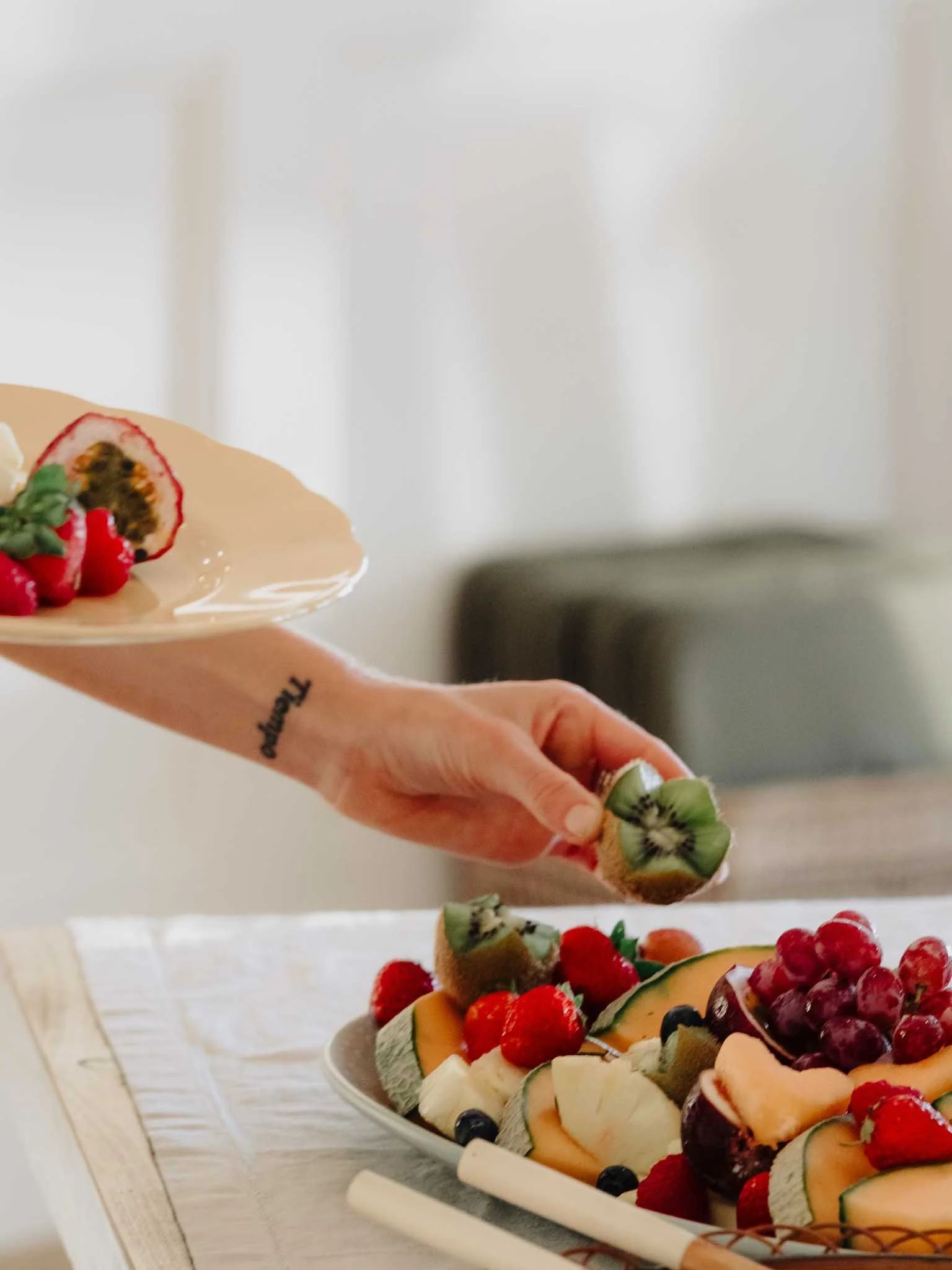 A person holding a sliced kiwi in front of a fruit platter containing various fresh fruits, including strawberries, blueberries, and melon.