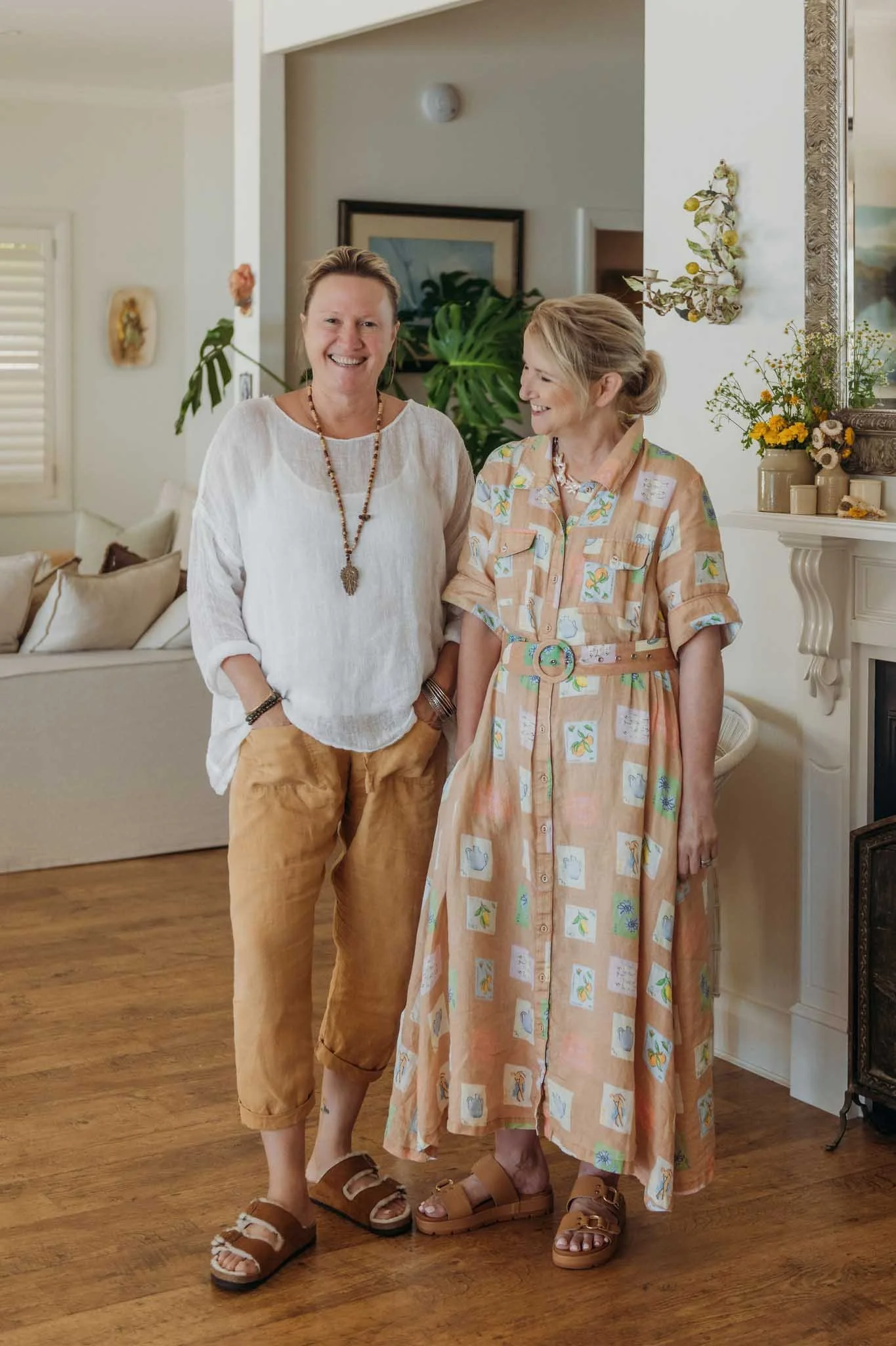 Two women standing indoors, smiling and looking at each other. One is wearing a white blouse and tan pants, the other in a patterned dress. They are wearing sandals.