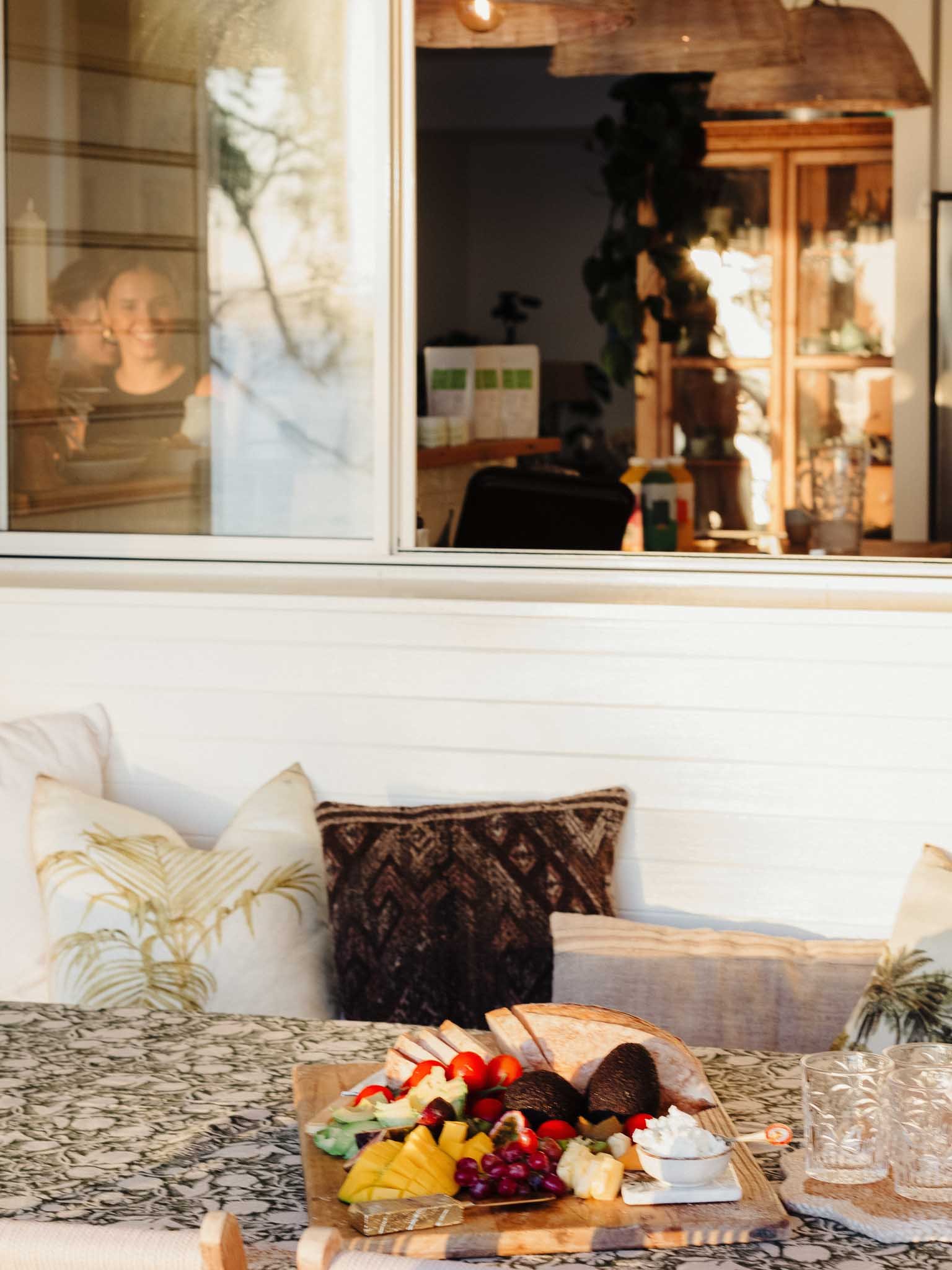 A wooden table with a platter of assorted fruit and black bread, with a room featuring decorative pillows on a sofa and glass of water in the background.