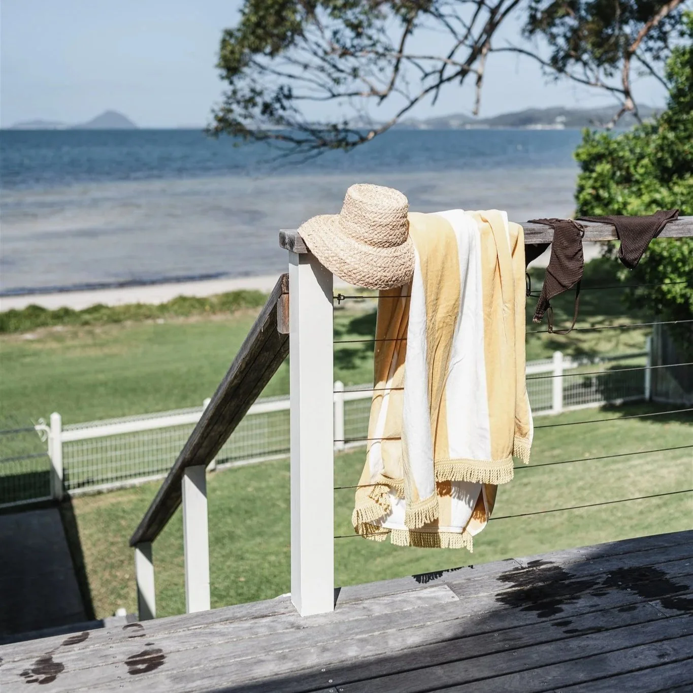A beach scene with clothes hanging on a railing, including a terry hat and towels, with a view of the ocean, trees, and distant landforms.