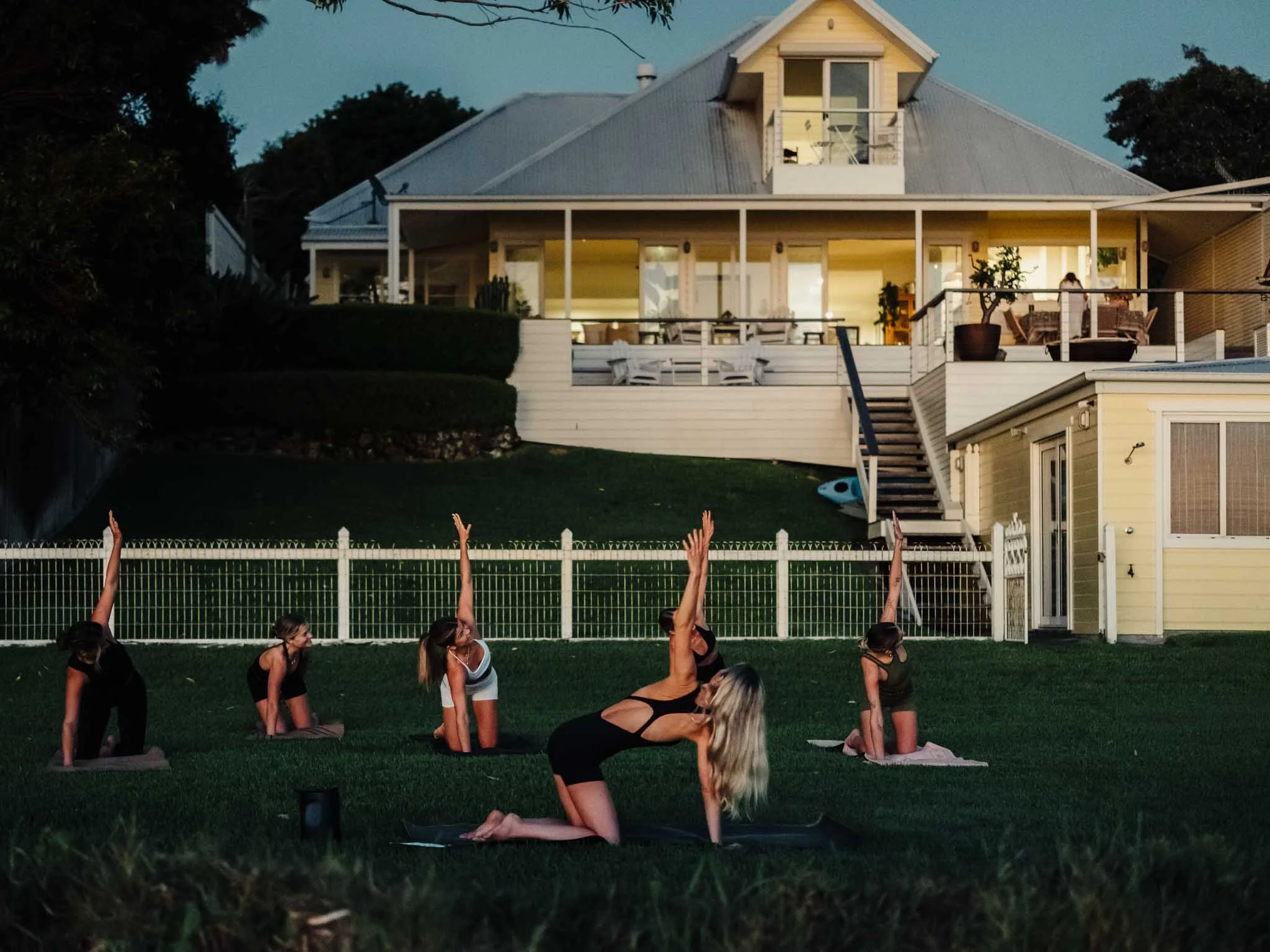 A group of people practicing yoga outdoors on a grassy lawn at sunset, facing a white house with a large balcony, trees, and a fence.