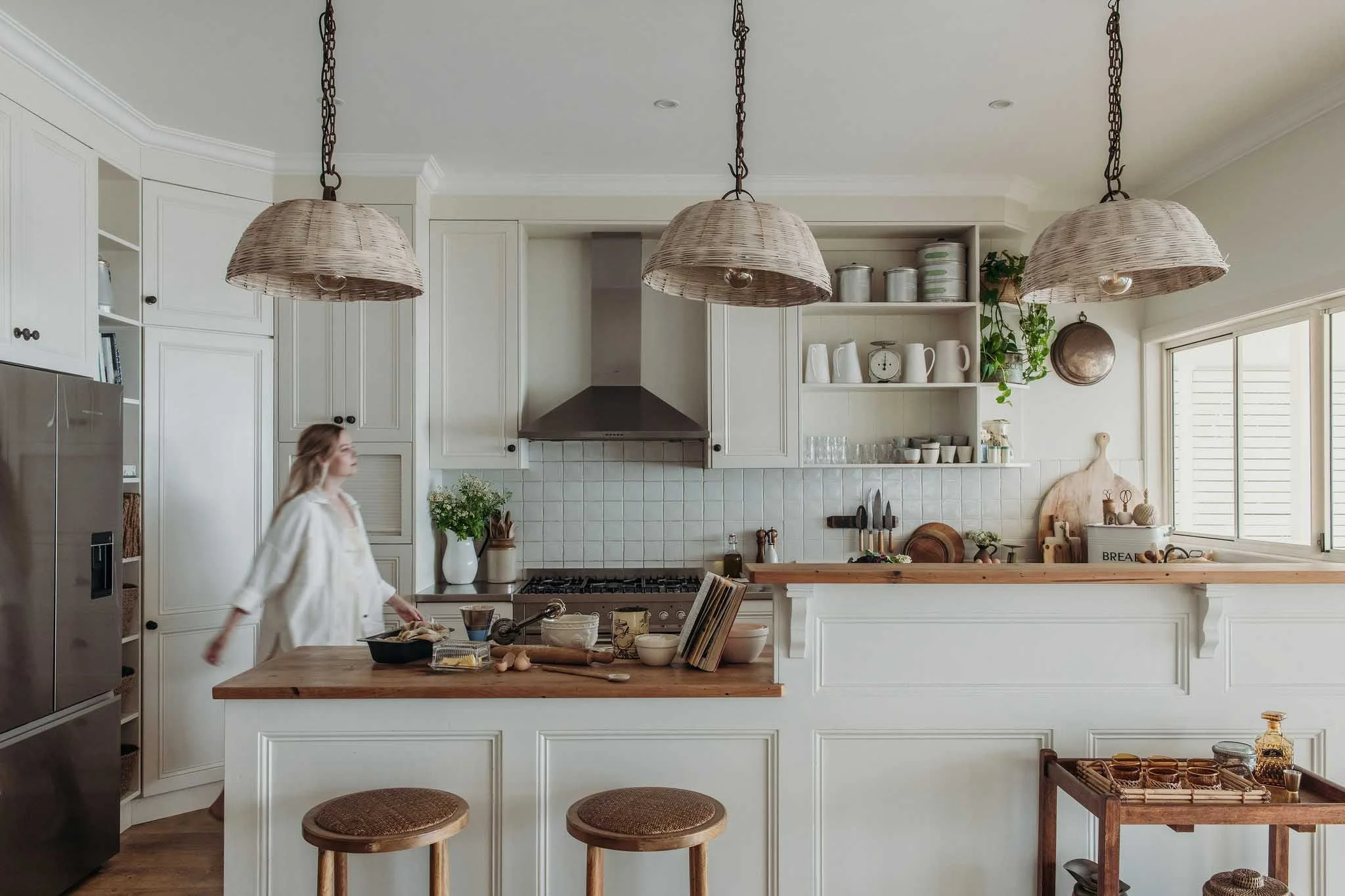 A kitchen with white cabinets, open shelving, and a central island. Three woven pendant lights hang above, and there is a person, possibly a young girl, walking in the foreground. The countertop has baking ingredients and utensils. There are two wove