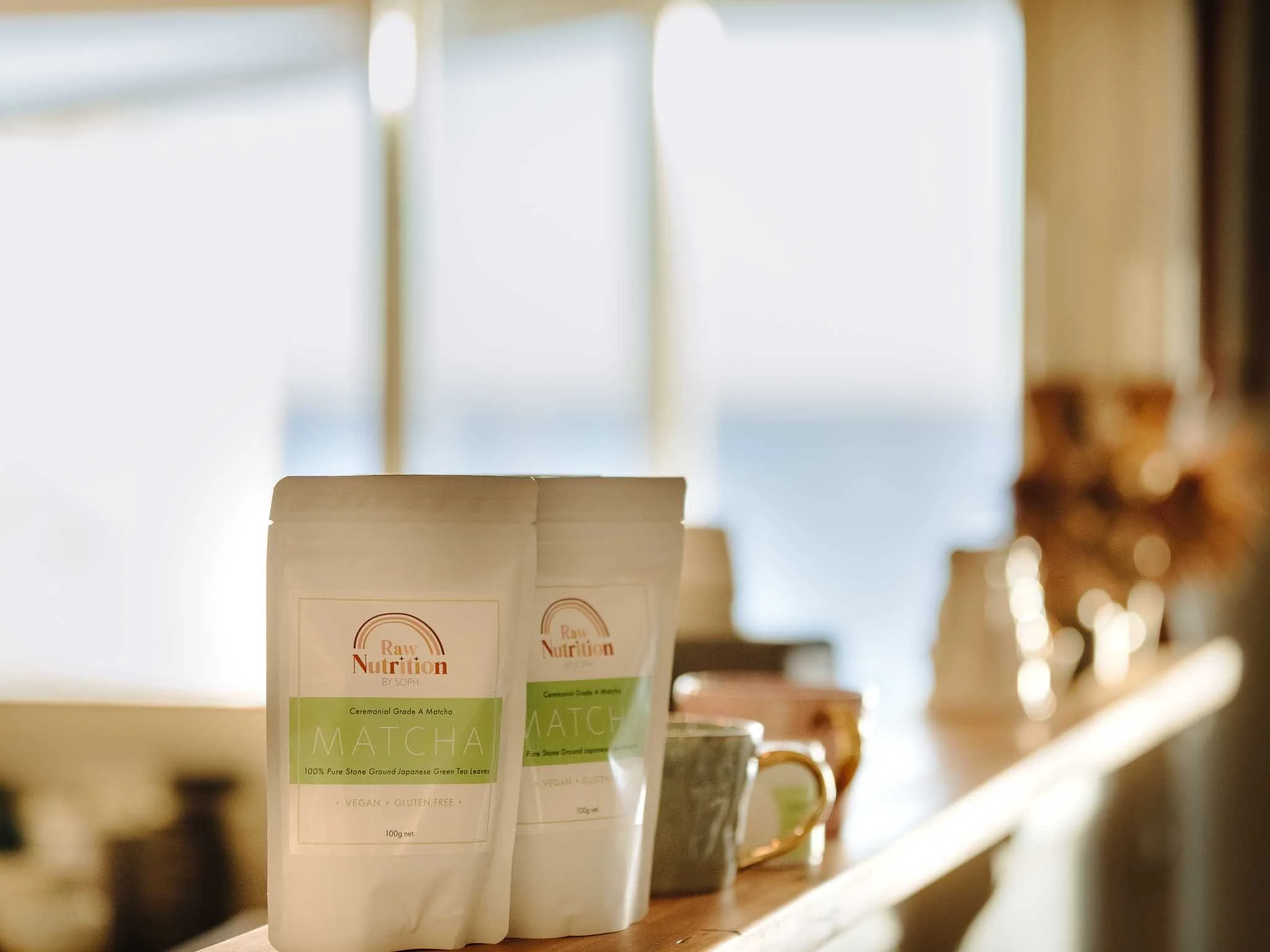 Close-up of white packets of matcha green tea powder with labels reading "Raw Nutrition" and "Ceremonial Grade A Matcha" on a wooden shelf in a store.