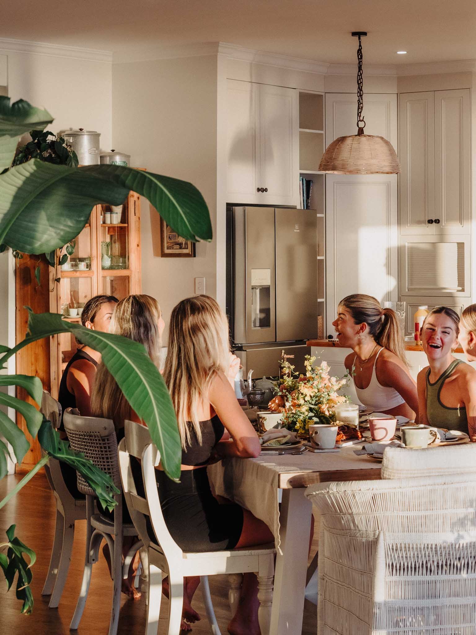 A group of women sitting around a dining table, smiling and talking, during a social gathering in a well-lit kitchen with white cabinets and a hanging wicker lamp.