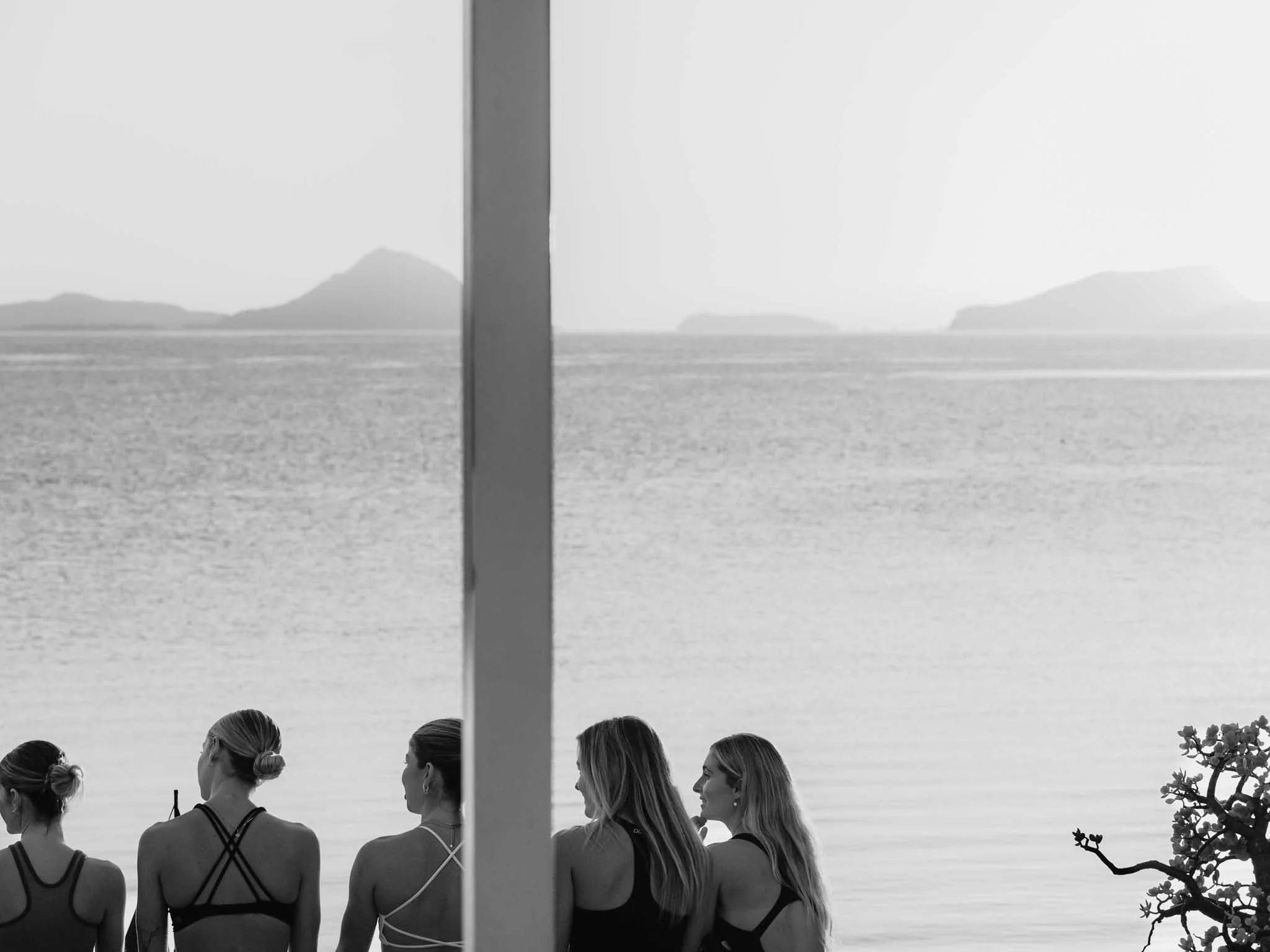 A black and white image of a group of women standing at the beach, facing the ocean, with distant islands in the background. The photograph is divided by a vertical pole in the center.