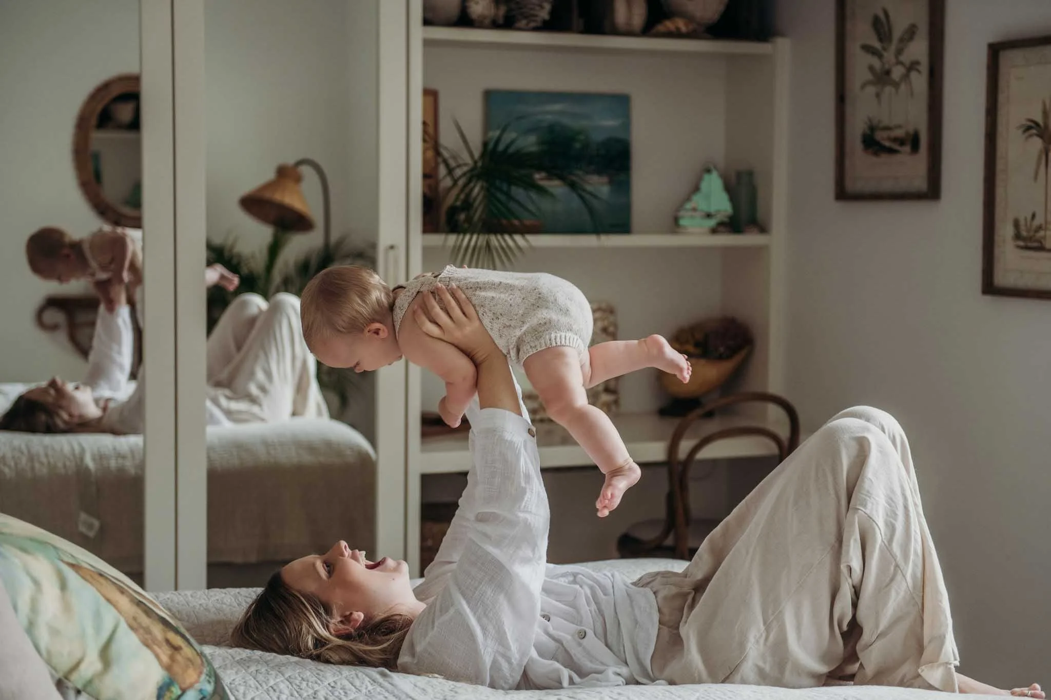 A woman lying on a bed holding a young boy upside down, both smiling and appearing to play. A mirror reflects the scene, and the room has warm, cozy decor with framed artwork, a desk lamp, and houseplants.