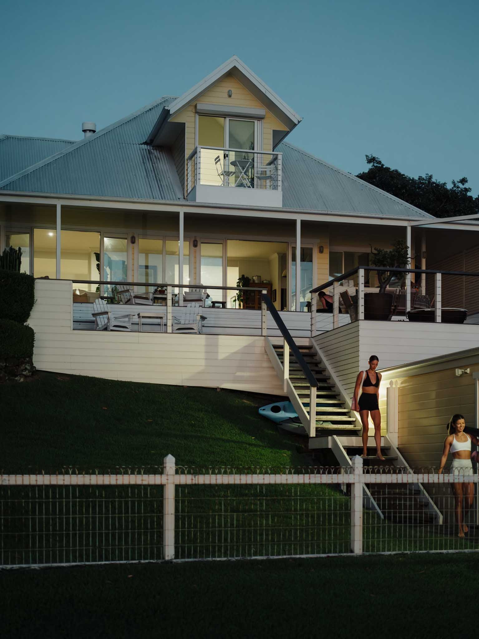 The image shows a two-story house with a metal roof and a balcony. The house has large glass doors leading to an outdoor patio with outdoor furniture. There are two women in sportswear walking down stairs outside the house, and a white fence surround