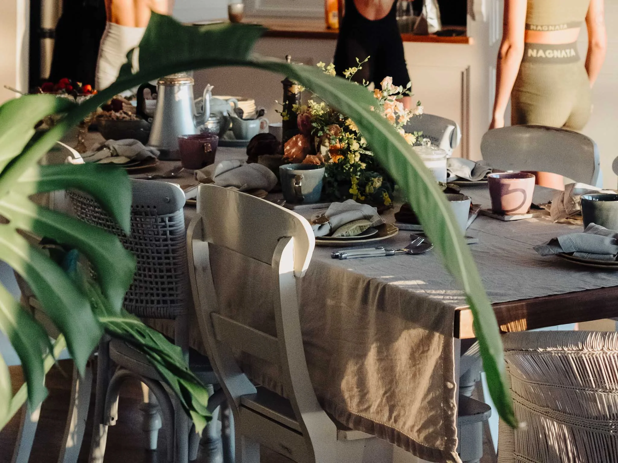 A dining table set for a meal with various plates, cups, utensils, and a floral centerpiece, with people standing around in a casual indoor setting.