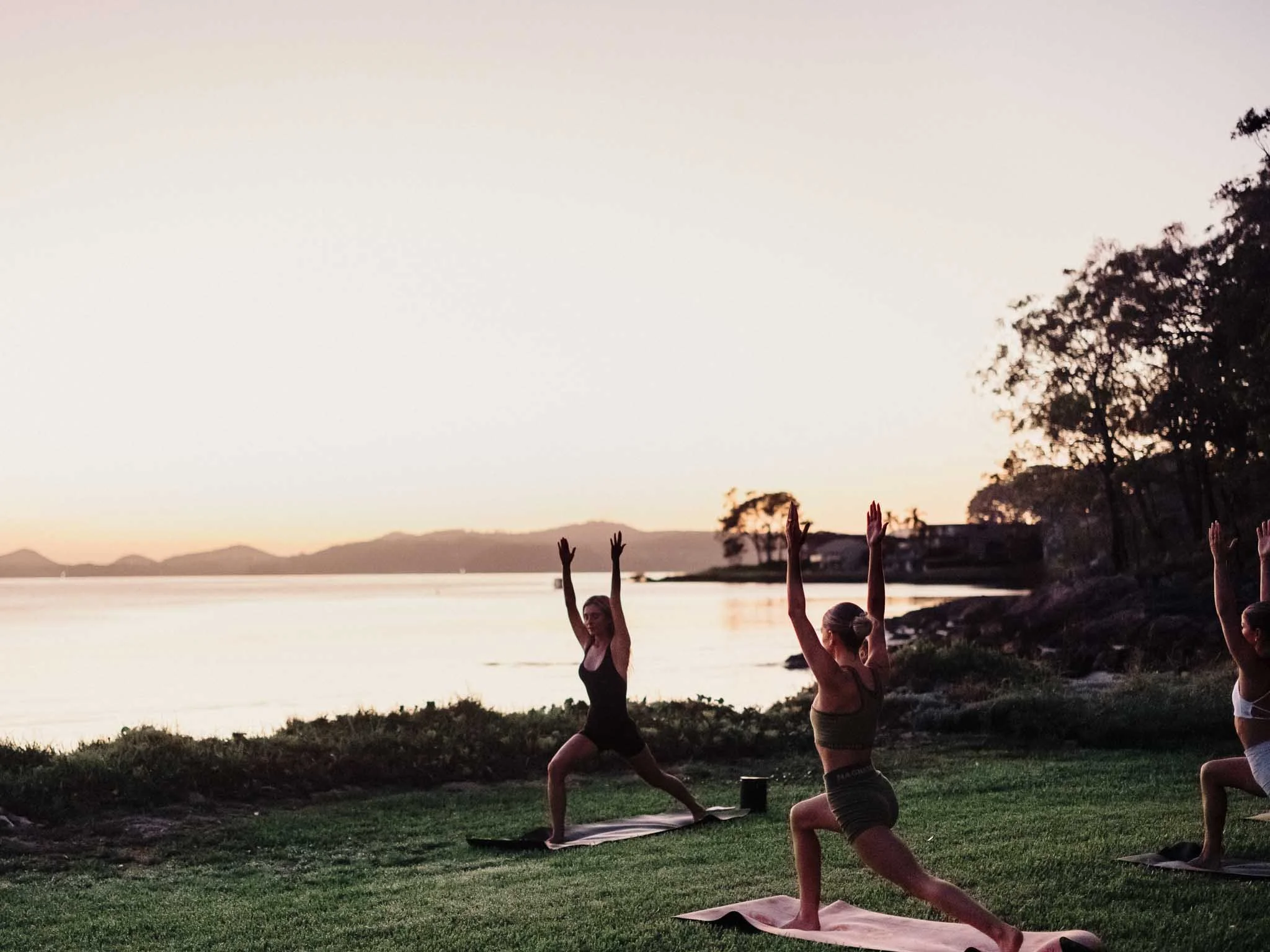 Group of people practicing yoga outdoors near a lake during sunset, some in Warrior pose, with trees and mountains in the background.