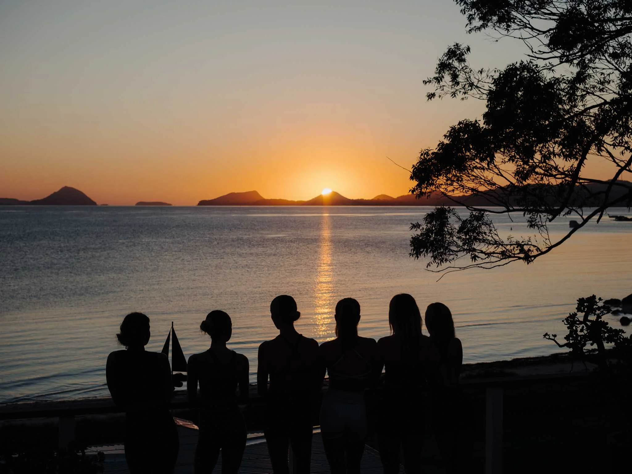 People sitting on the beach watching the sunset over the water with small islands in the distance and trees in the foreground.