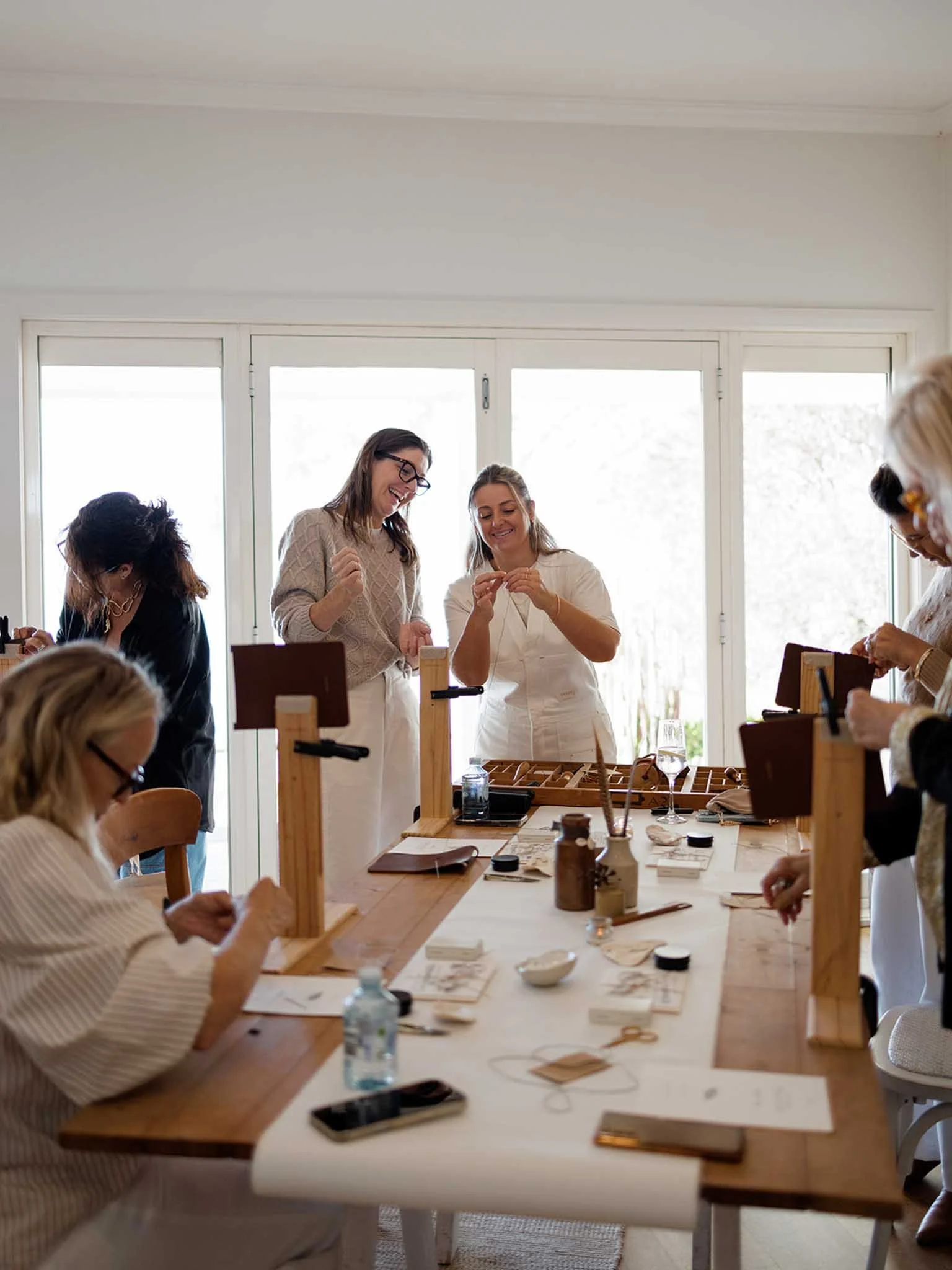 Women participating in a craft workshop in a bright room with large windows, working on jewelry or similar crafts at a long table.