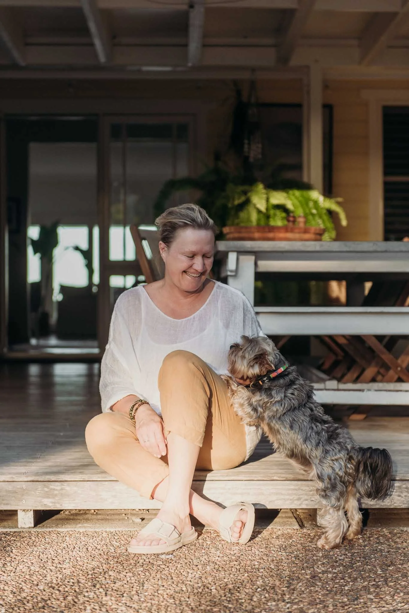 A woman sitting on a wooden porch, smiling and playing with her dog, a small furry breed, in warm natural light.