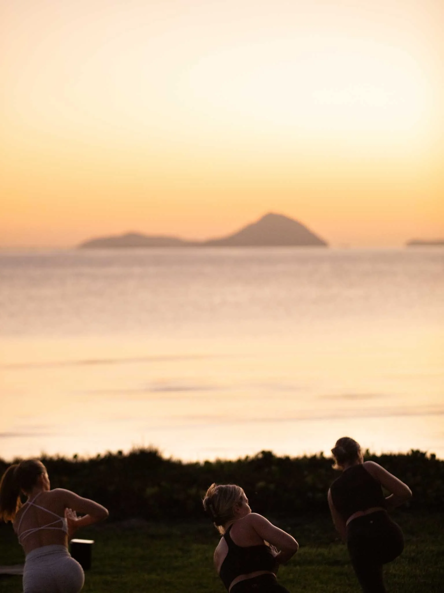People practicing yoga outdoors during sunset by the water with islands in the background.