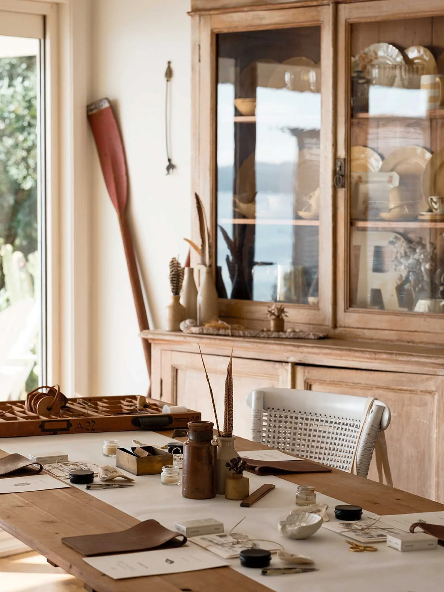 An art studio workspace with a large wooden table, art supplies, sketches, and a glass-front wooden cabinet displaying artwork and decorative objects. Natural light comes through a nearby window.