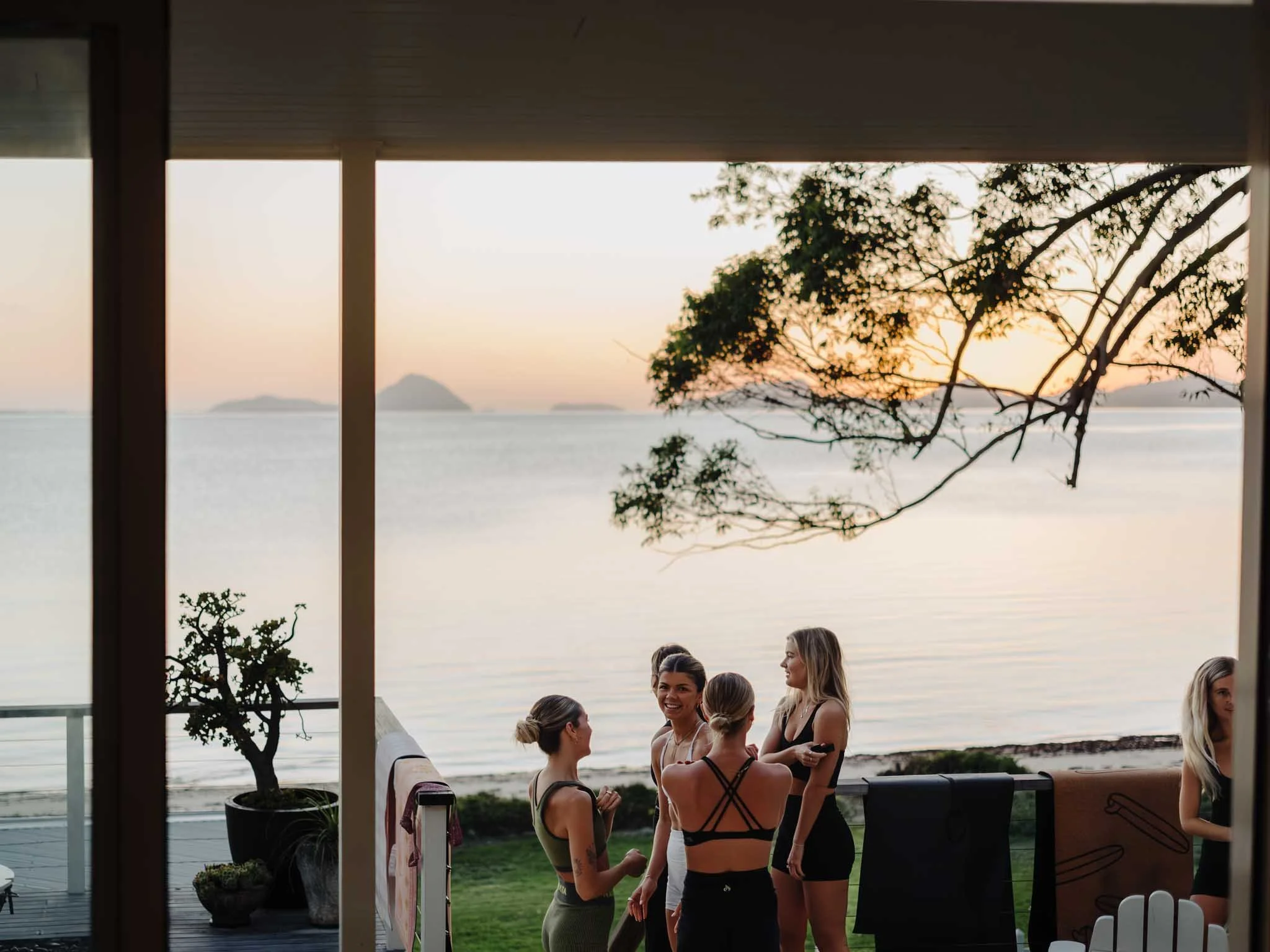 Group of women socializing on a terrace overlooking the ocean during sunset, with trees and islands in the background.