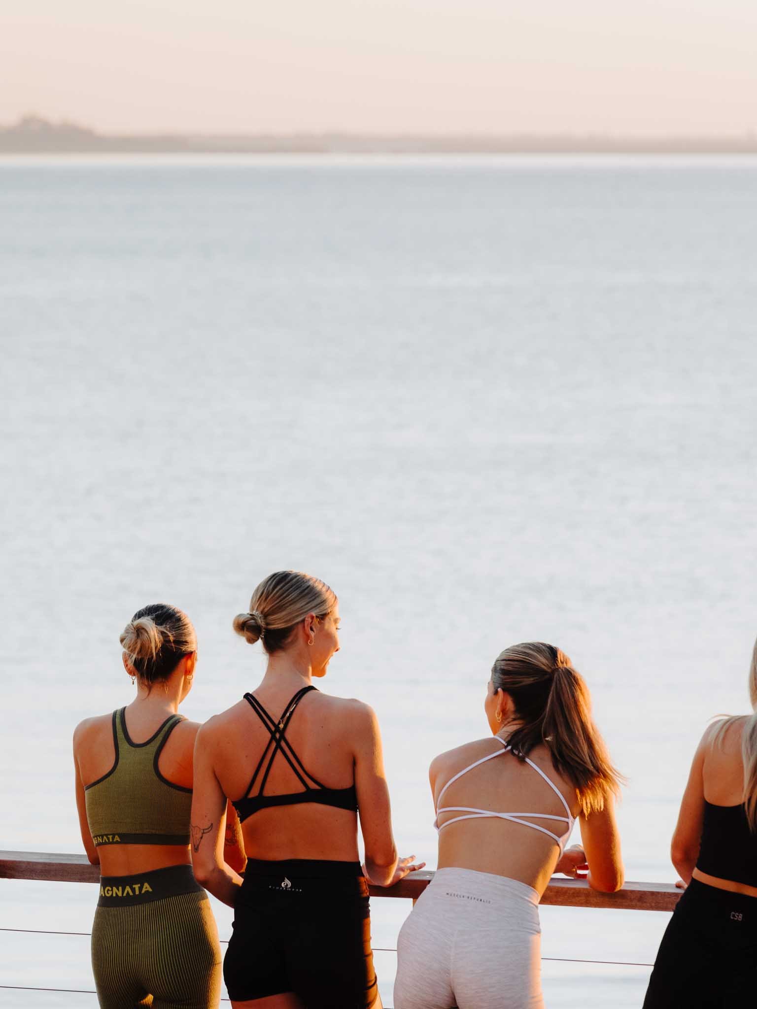 A group of women with their hair tied back, looking out at water during sunset.