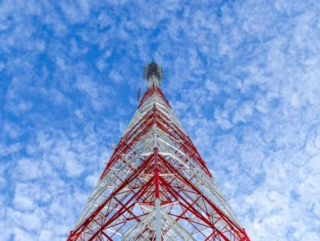 A tall communication tower with red and white structural elements reaching up into a partly cloudy blue sky.
