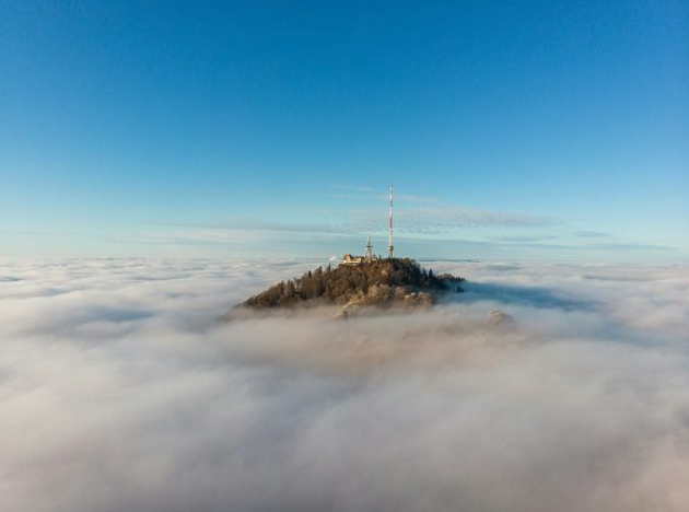 A mountain topped with trees and a communication tower, surrounded by a sea of clouds with a clear blue sky.