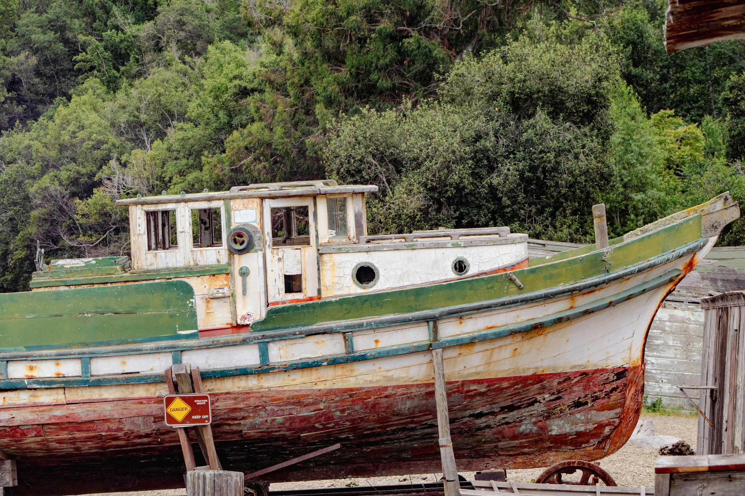 An old, abandoned boat with peeling and faded paint, resting on land surrounded by trees, with a warning sign indicating danger.