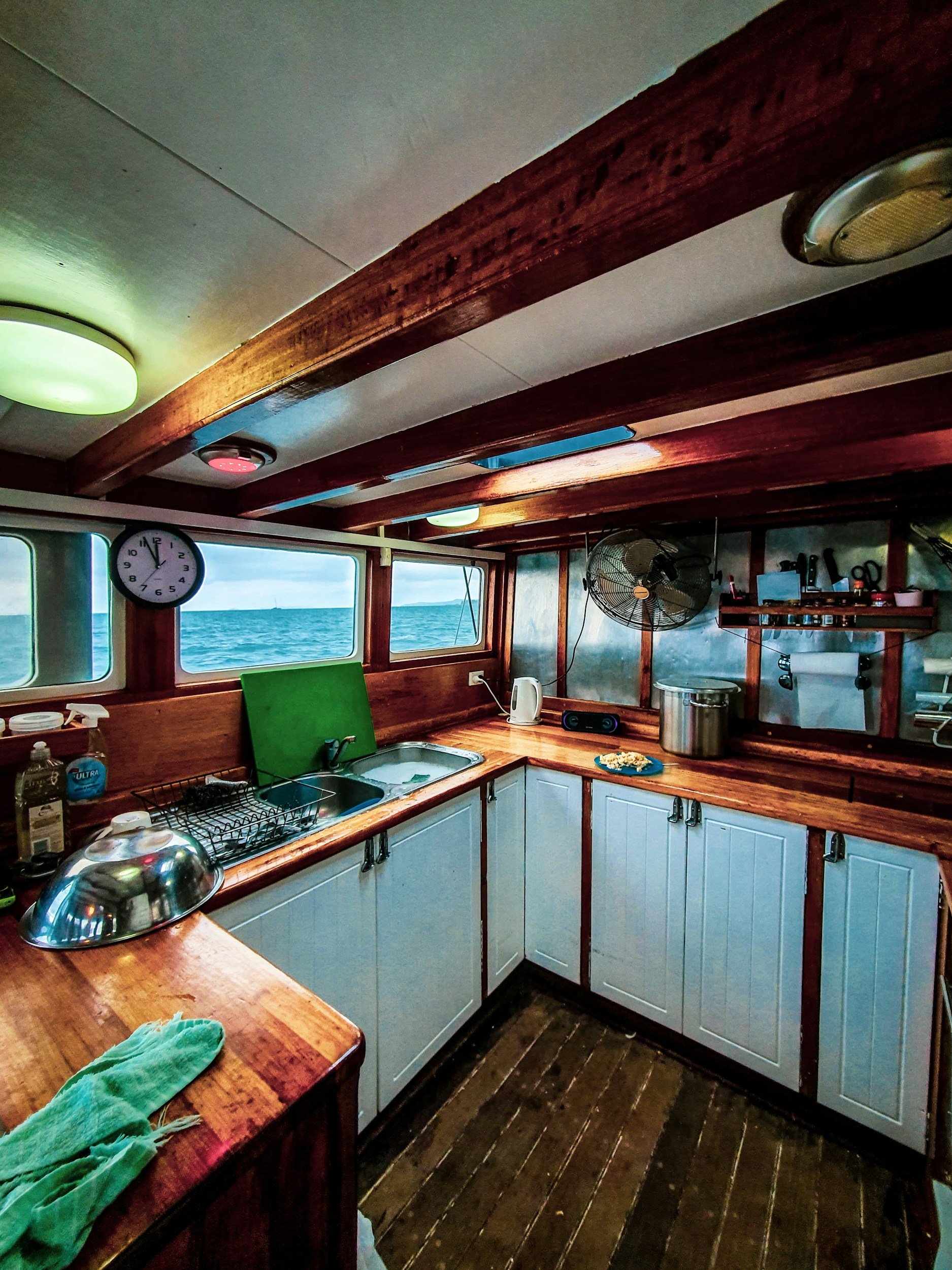 Cozy kitchen space with wooden countertops and cabinets, with windows showing a view of the sea, ceiling lights, a clock, and various kitchen items.