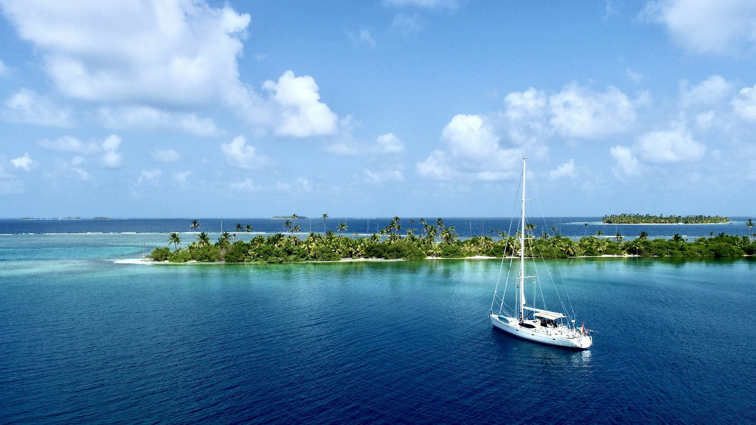 A sailboat floating on clear blue water near a tropical island with lush greenery, palm trees, and small islands in the background under a partly cloudy sky.