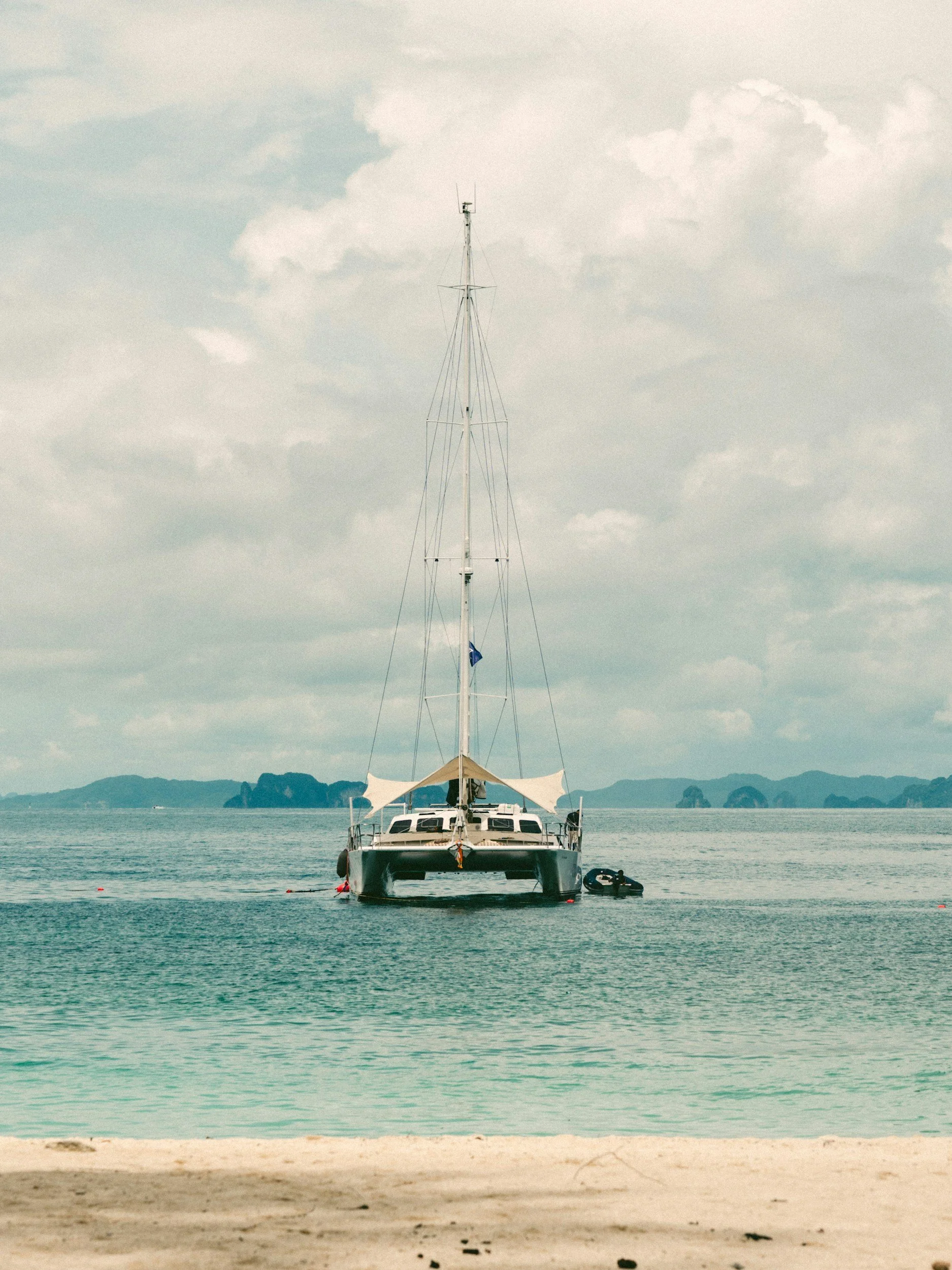 A yacht anchored in calm ocean water near a sandy beach with distant islands and a cloudy sky.