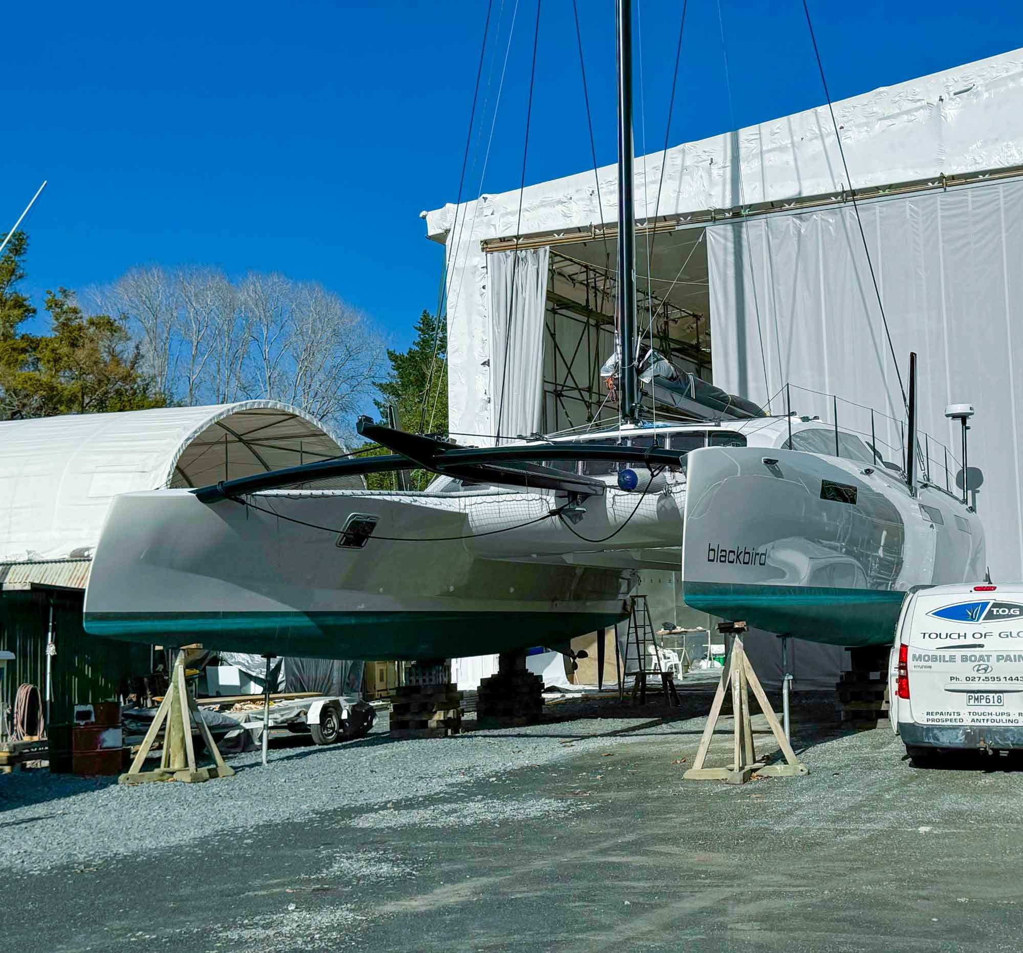 The image shows a boatyard with two boats on trailers under a protective canopy, with a white building and some equipment or supplies around, and trees in the background against a clear blue sky.