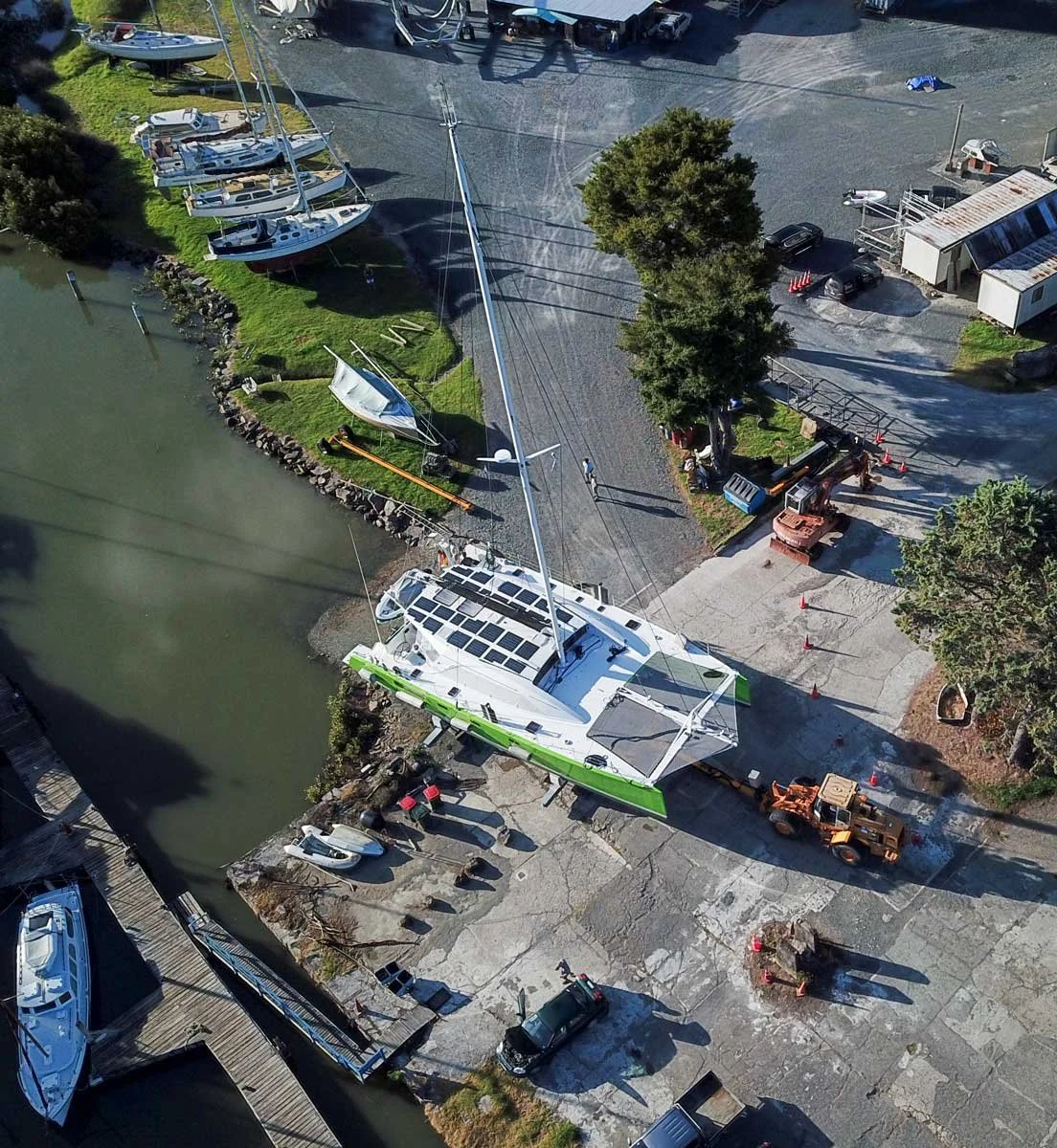 An aerial view of a marina with a large sailboat being lifted out of the water by a vehicle, surrounded by smaller boats, trees, parked cars, and industrial equipment.