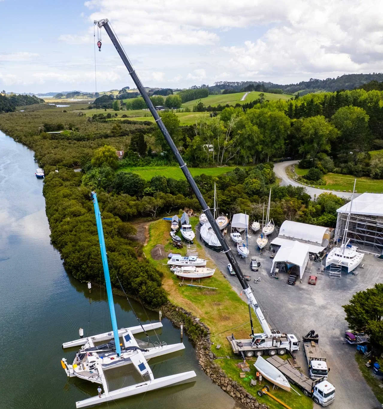 Aerial view of a boatyard with boats on land and water, a crane lifting a boat, and green landscape in the background