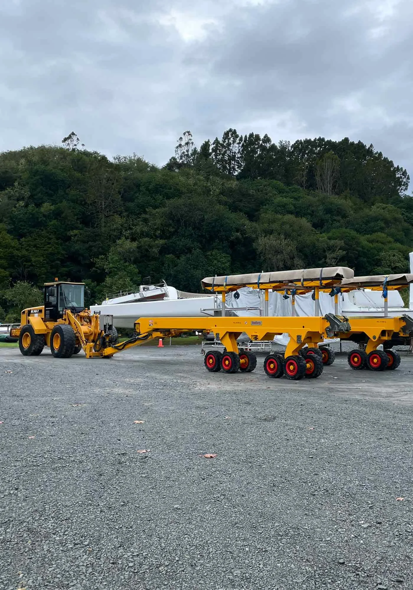 A yellow construction vehicle lifting a boat with a white cover on a gravel lot. In the background, there are trees and overcast sky.